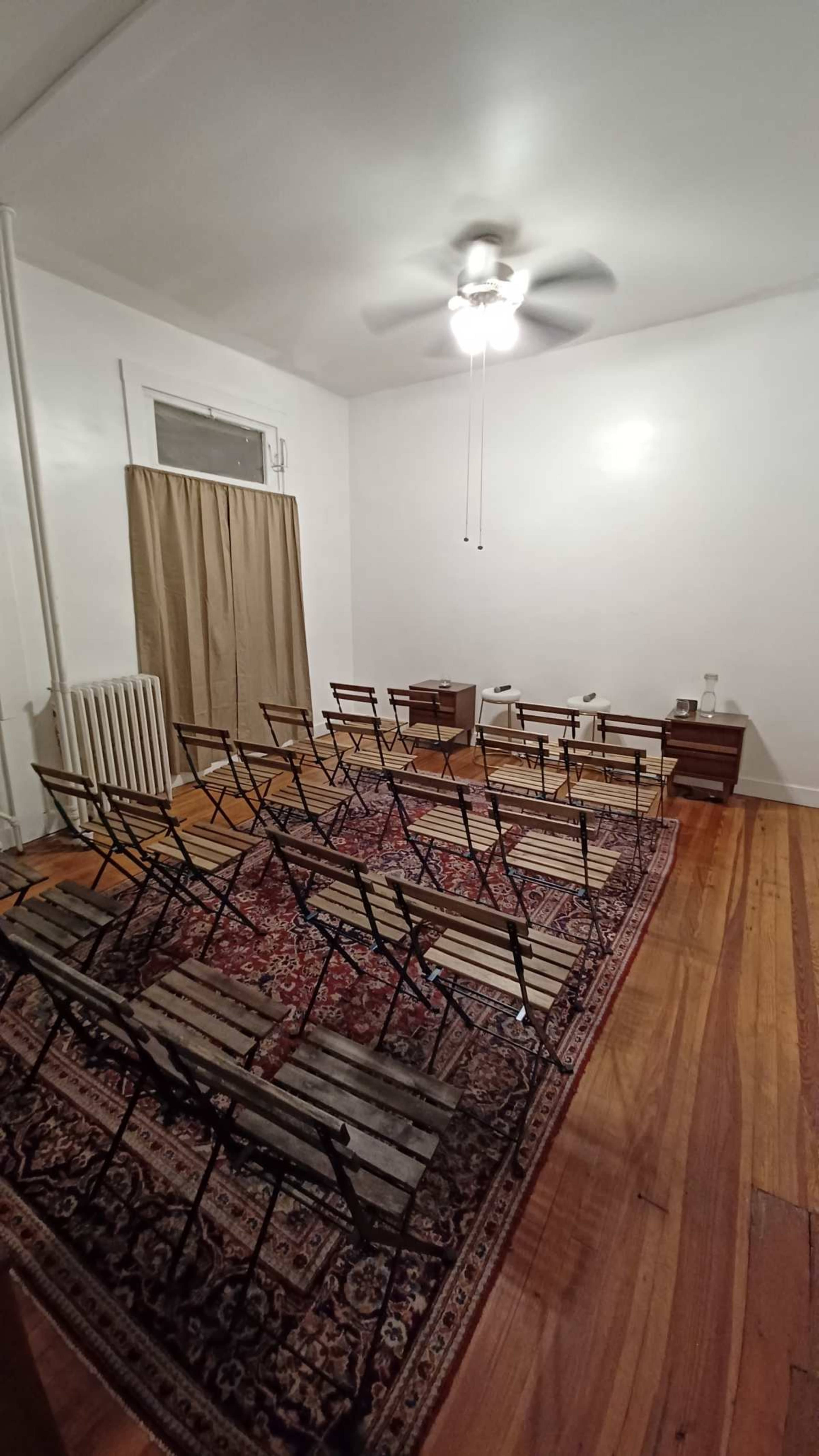 The image shows a sparsely furnished room with several folding chairs arranged on a patterned rug, a curtain covering a window, and a ceiling fan.