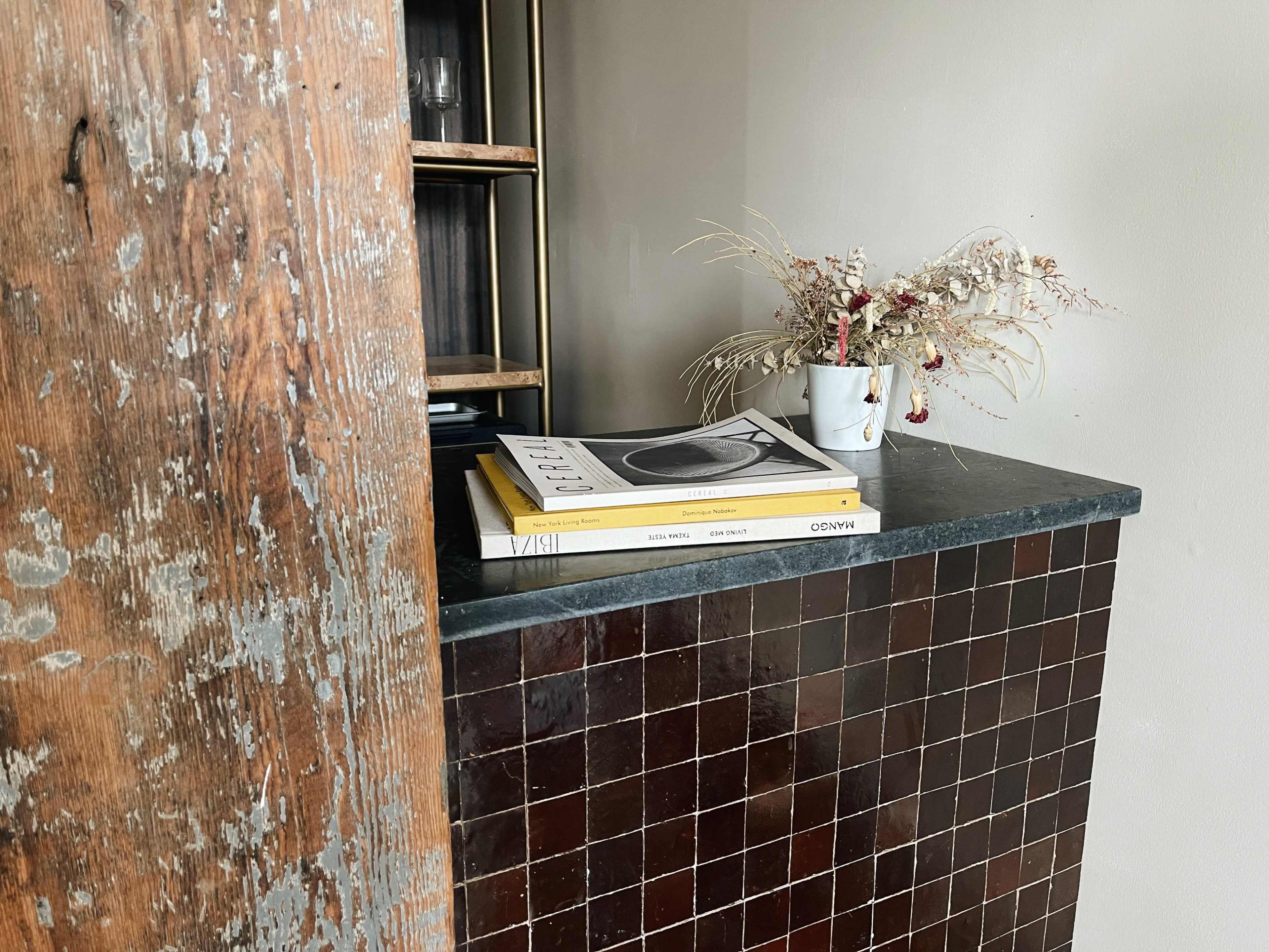 A wooden beam frames a countertop with stacked books and a small vase of dried flowers against a neutral wall.