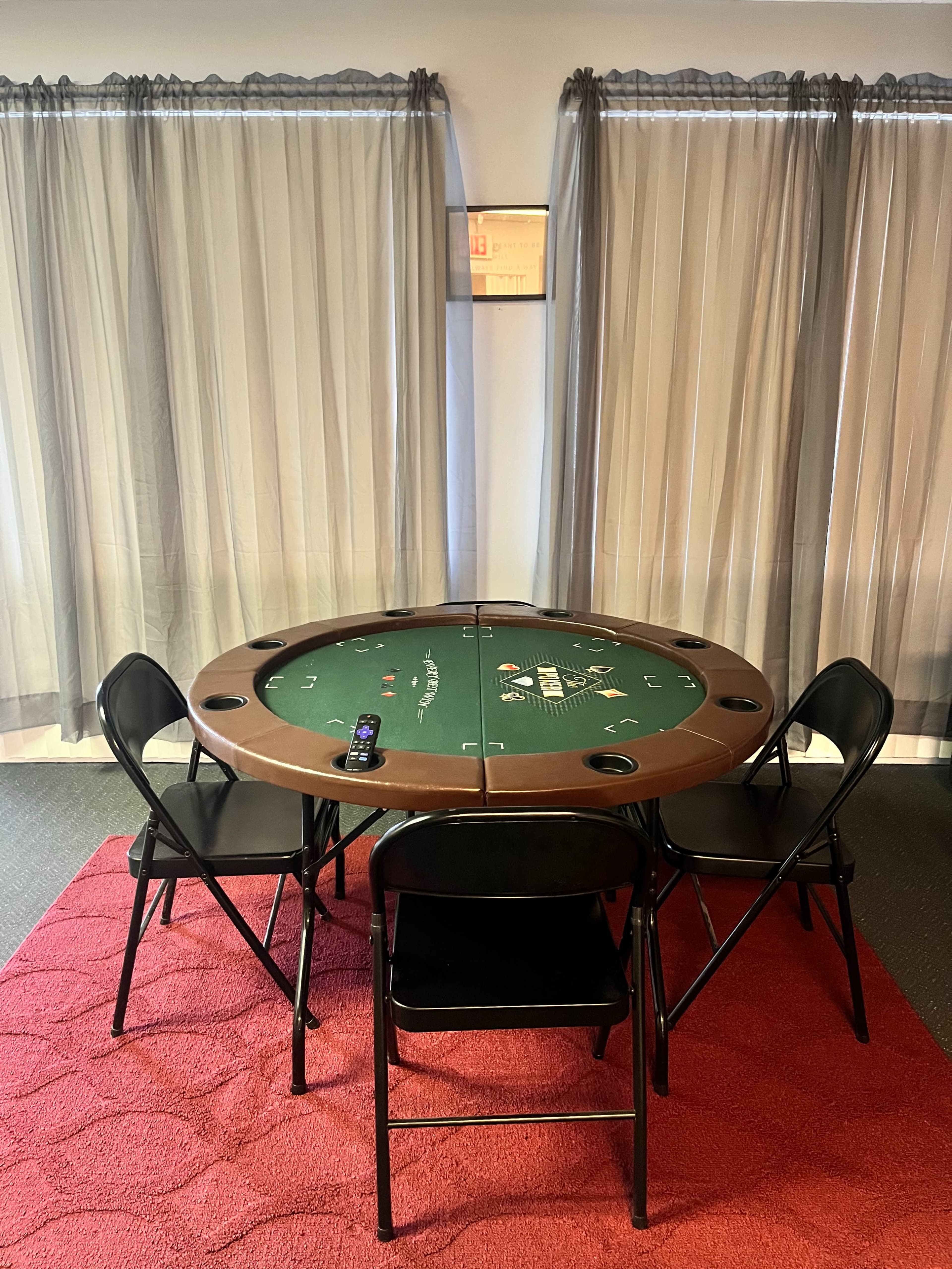 A round poker table surrounded by four black folding chairs is set up on a red carpet in a room with sheer curtains.