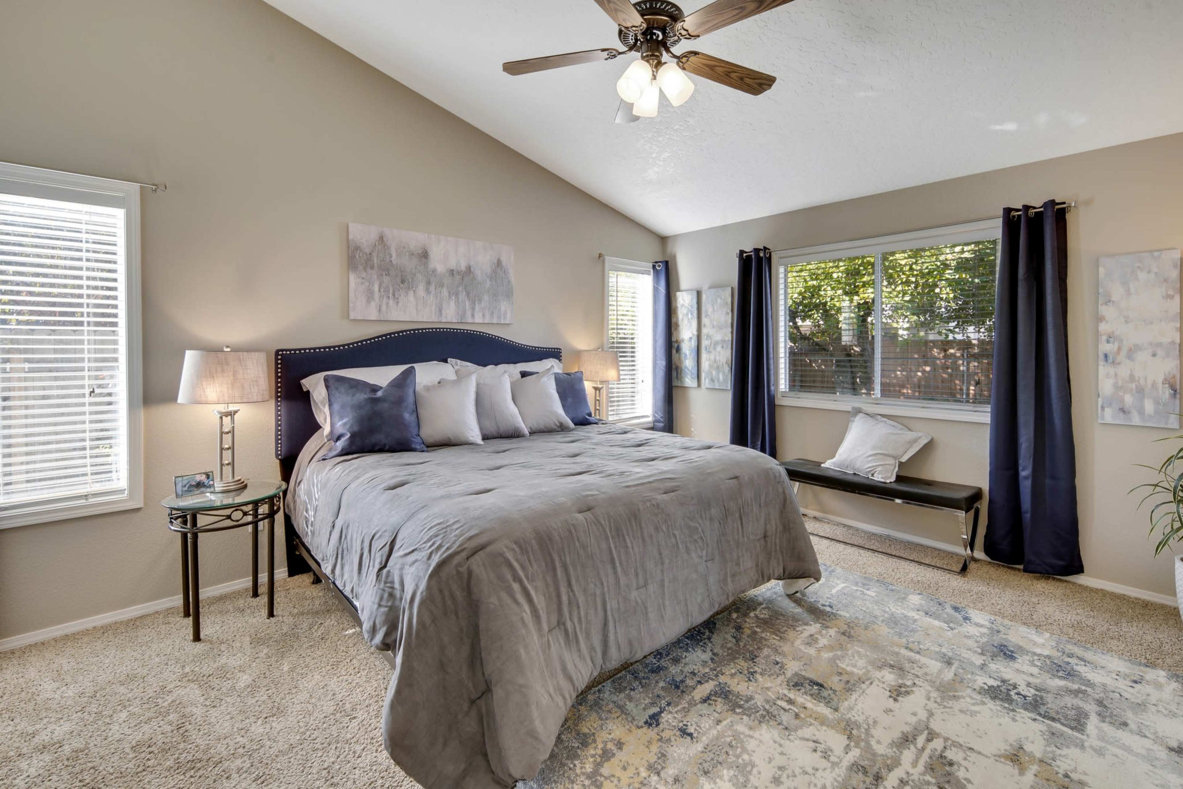 A well-lit bedroom featuring a large bed with decorative pillows, a bedside table with a lamp, and a window overlooking a garden.