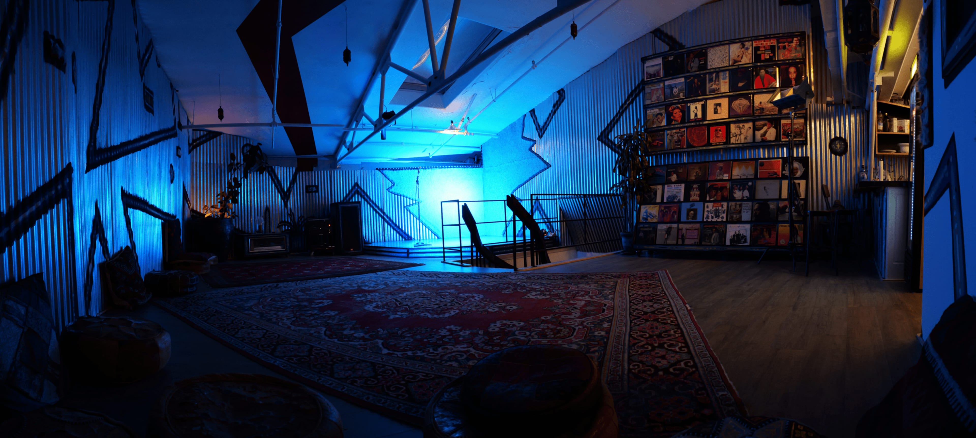 A spacious room with blue lighting, featuring a wall of vinyl records, a patterned rug, and a staircase leading to a lower level.