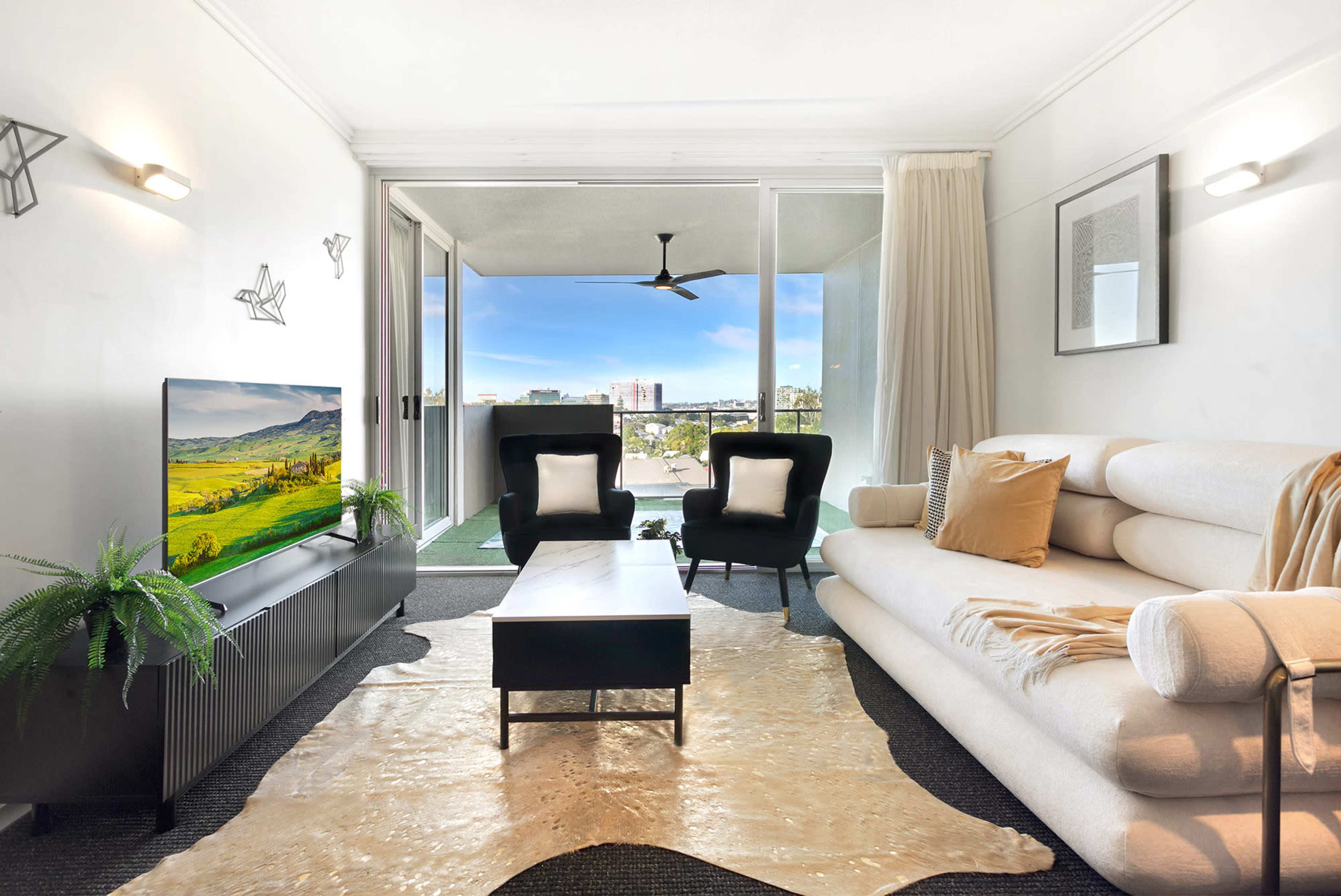 A modern living room features a light-colored sofa, two black armchairs, a coffee table, and a television, with large windows offering a view of the outdoors.