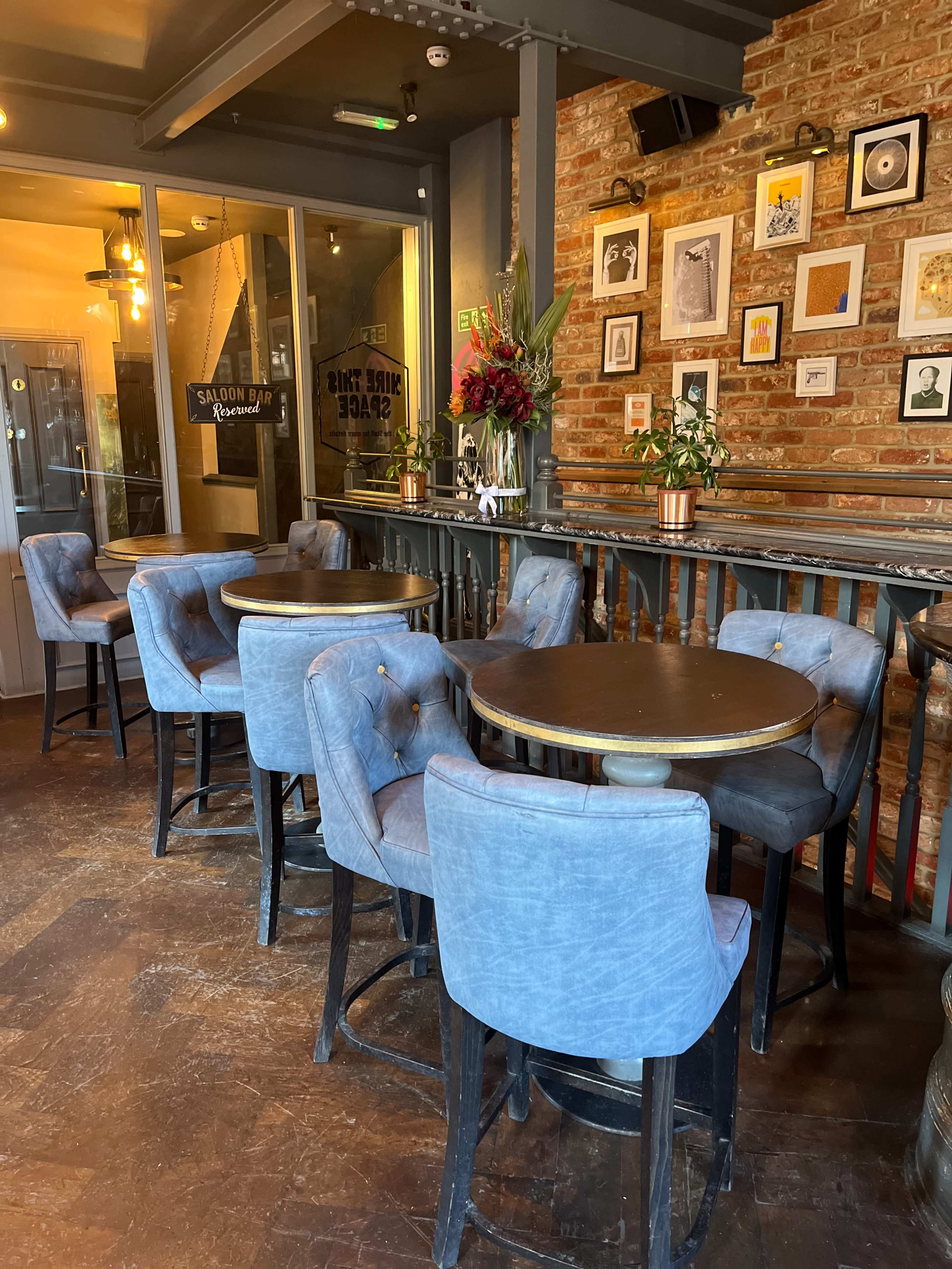 The image shows a cozy dining area featuring several round tables surrounded by upholstered chairs, with a brick wall adorned with framed artwork in the background.