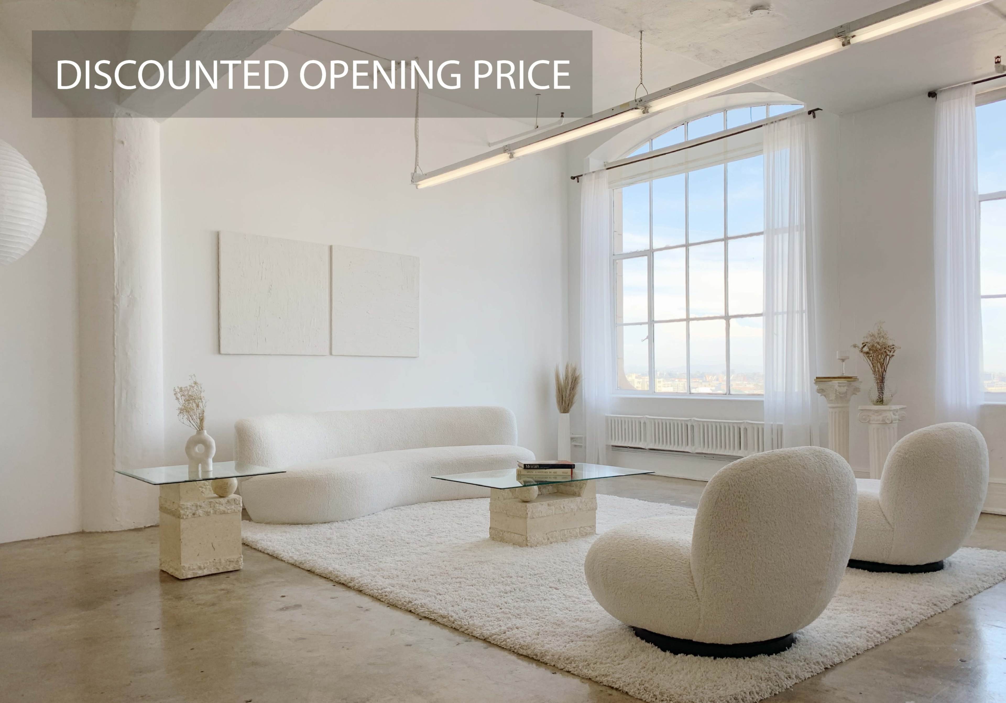 A spacious, minimalist living room featuring a white, plush sofa, two rounded chairs, and a glass coffee table, all set against a backdrop of large windows.