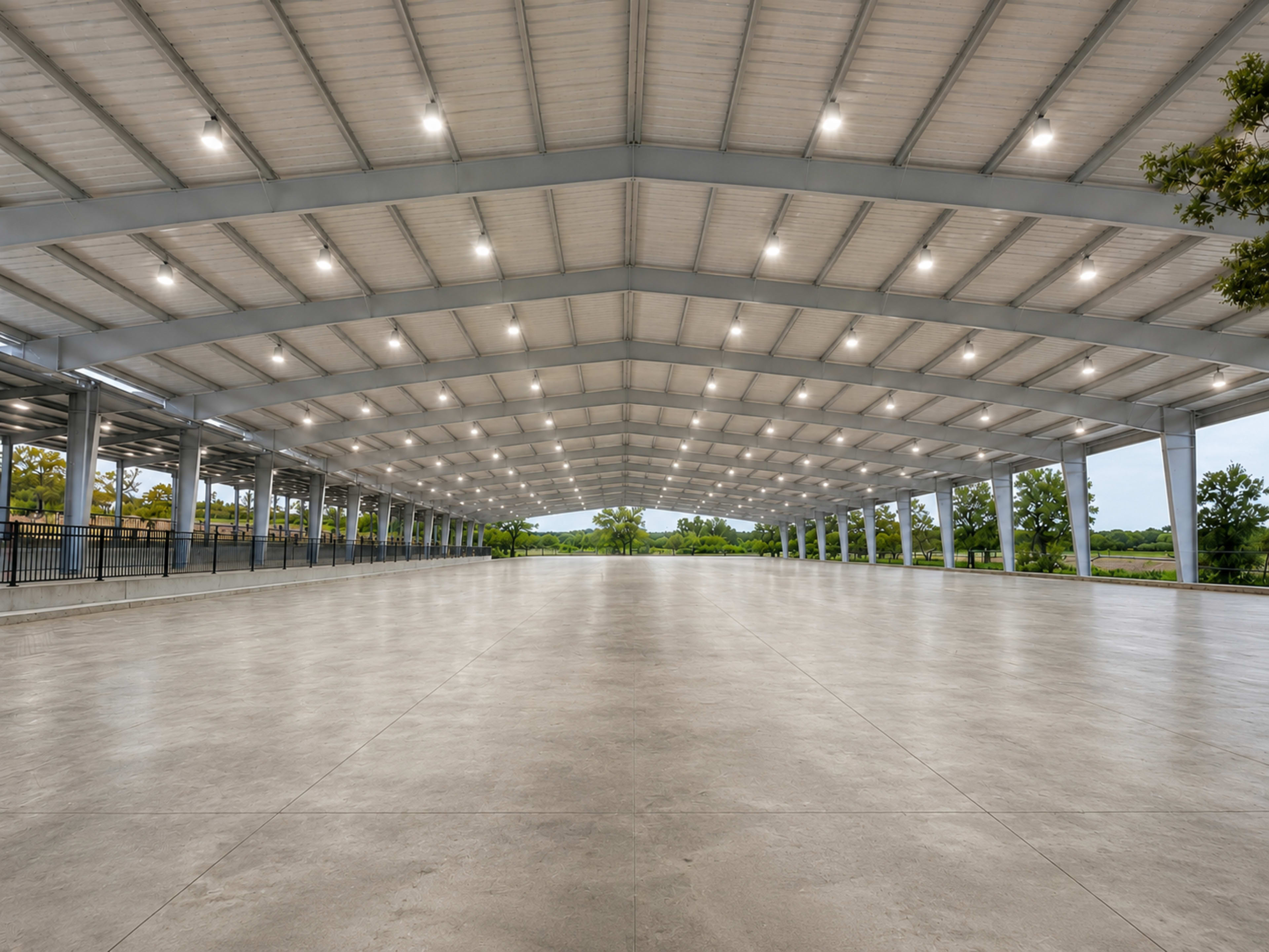 The image shows a large, empty indoor space with a high, metal-framed ceiling and concrete flooring, surrounded by green trees in the background.