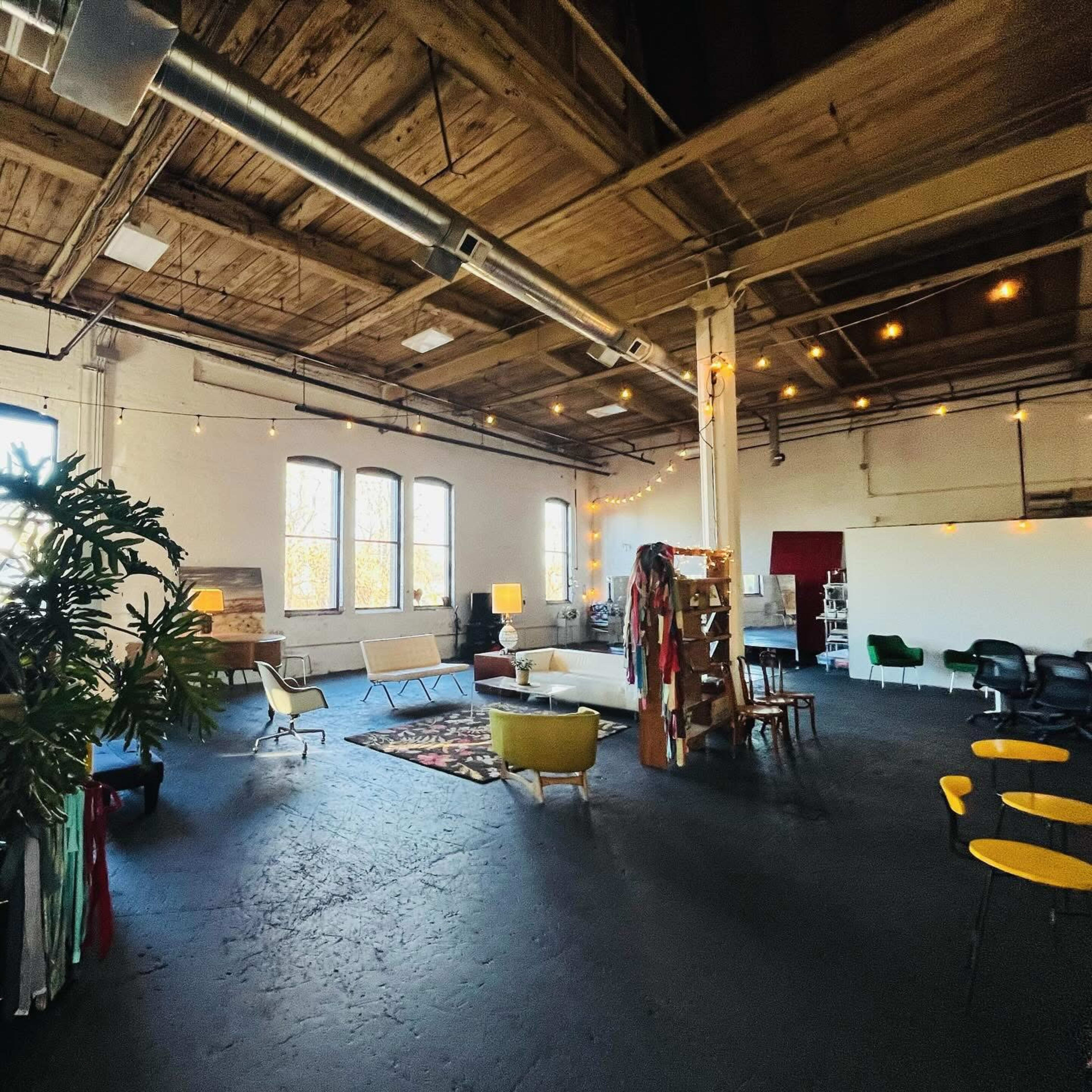 The image shows a spacious, well-lit interior with wooden beams, multiple seating areas, and decorative plants, set up for a creative workspace or gathering.