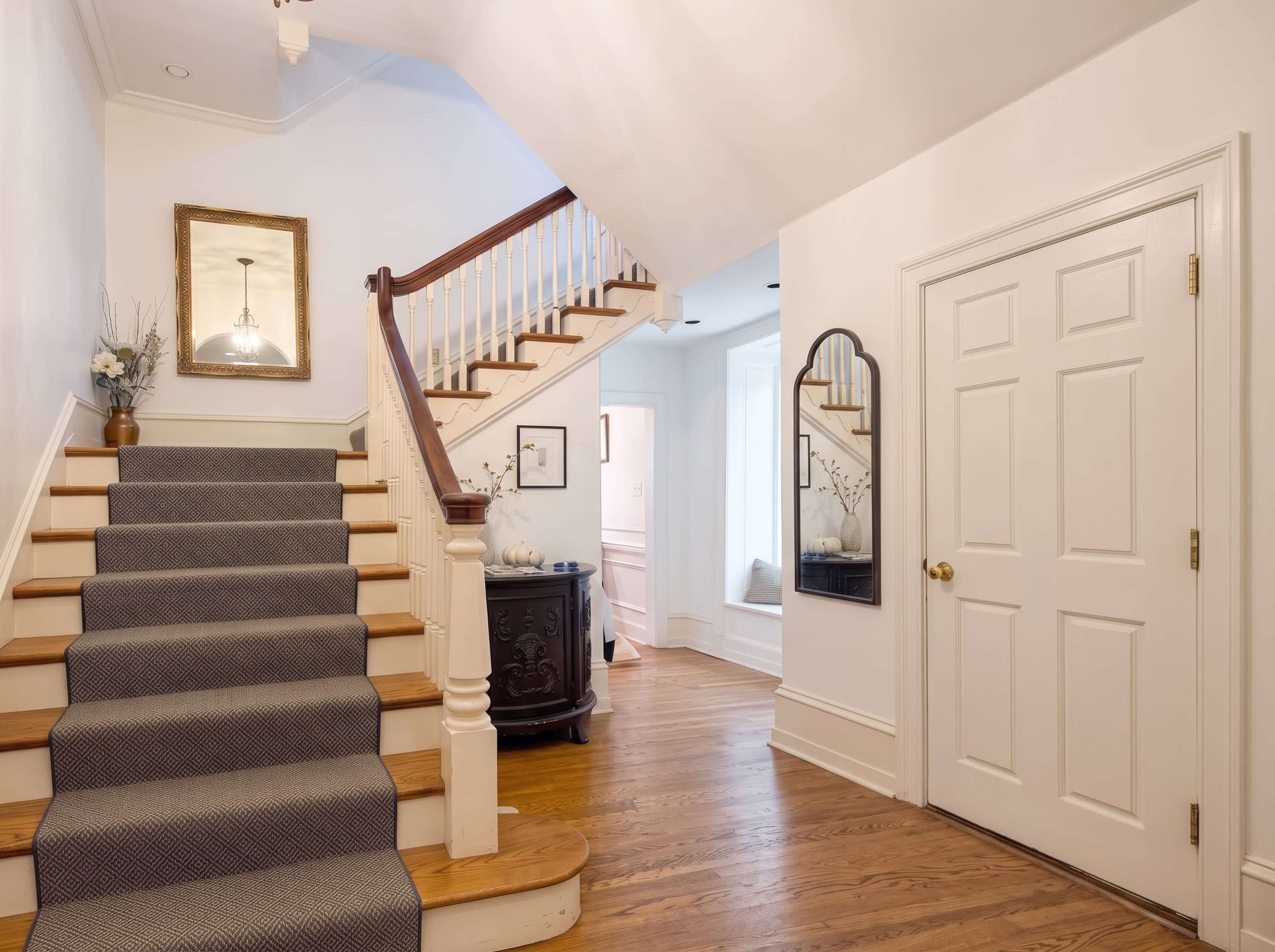 A well-lit foyer features a wooden staircase with a carpet runner, a decorative mirror, and a dark antique cabinet against the wall.