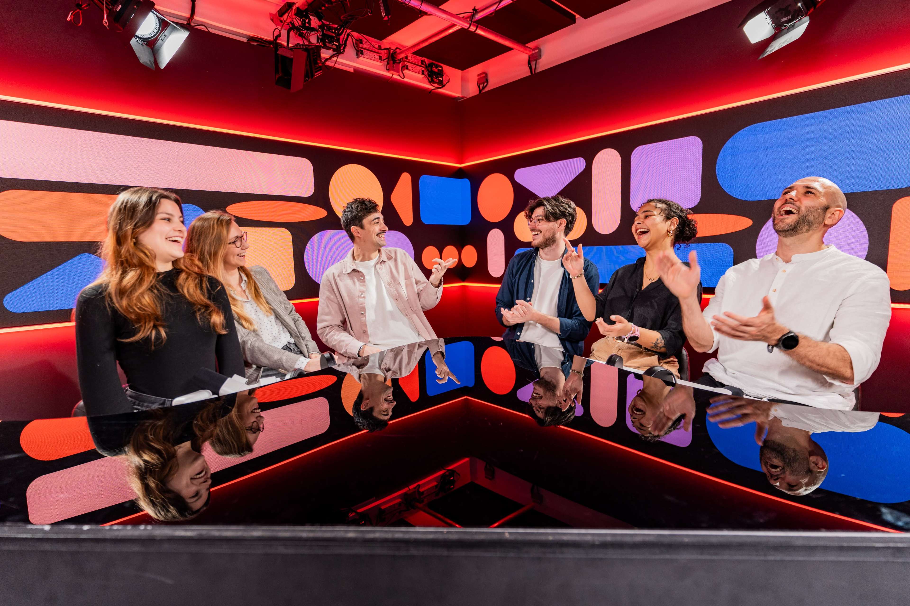 A group of six people sits around a reflective table in a brightly colored studio, sharing laughter and conversation.