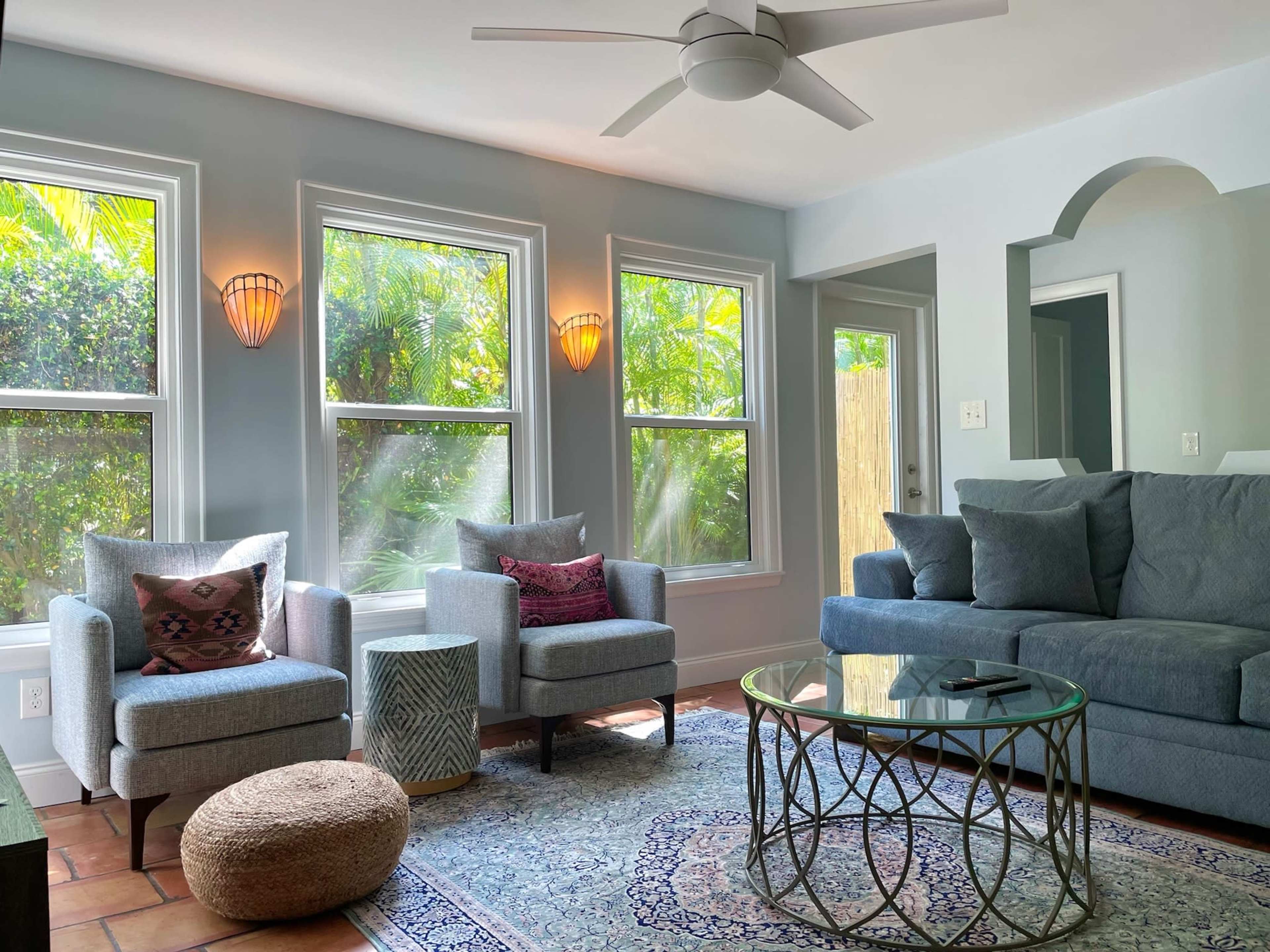 The living room features two gray armchairs, a glass coffee table on a patterned rug, and large windows allowing natural light to illuminate the space.