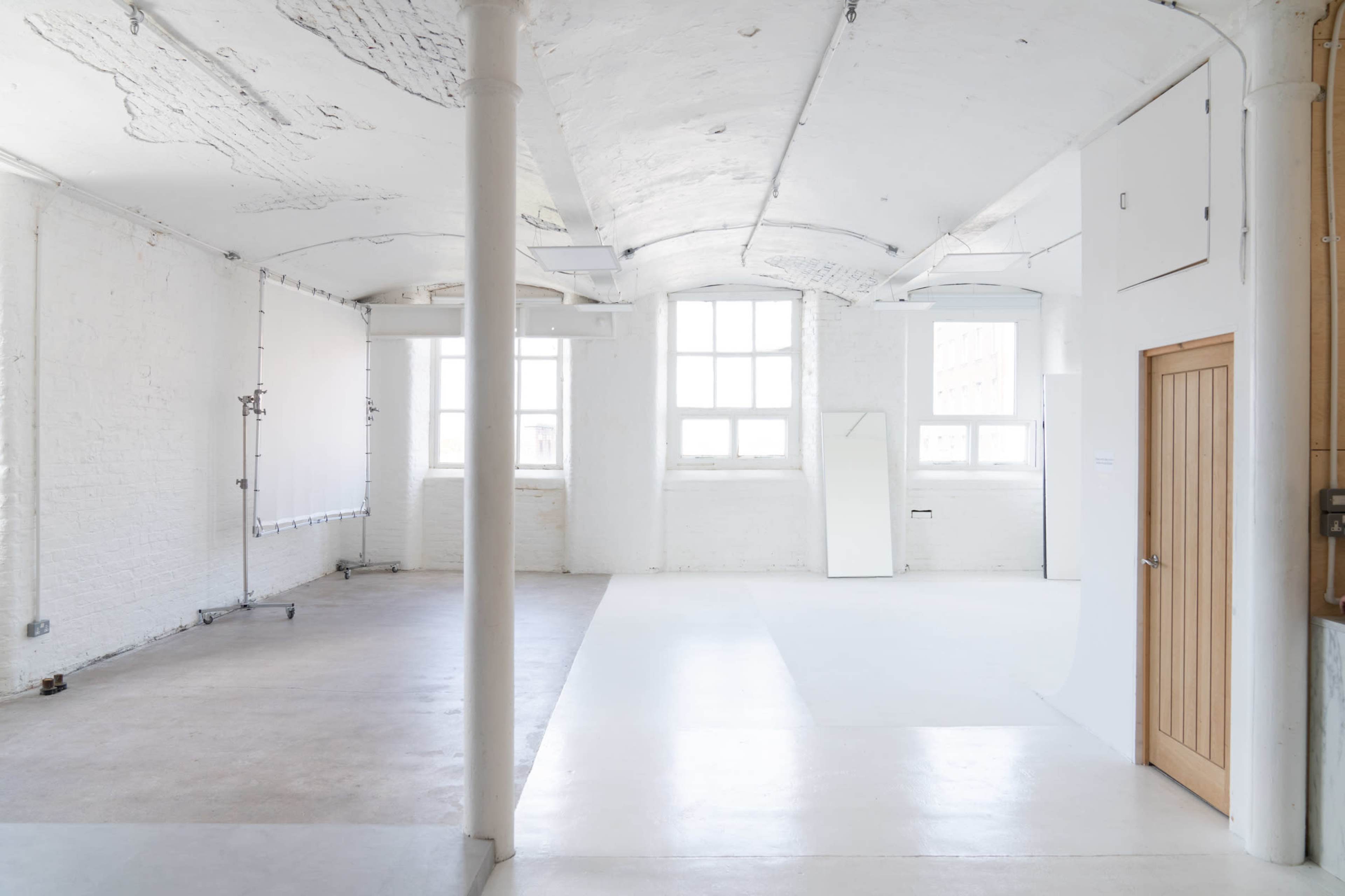A bright, spacious studio with white walls, large windows, and a seamless backdrop on one side.