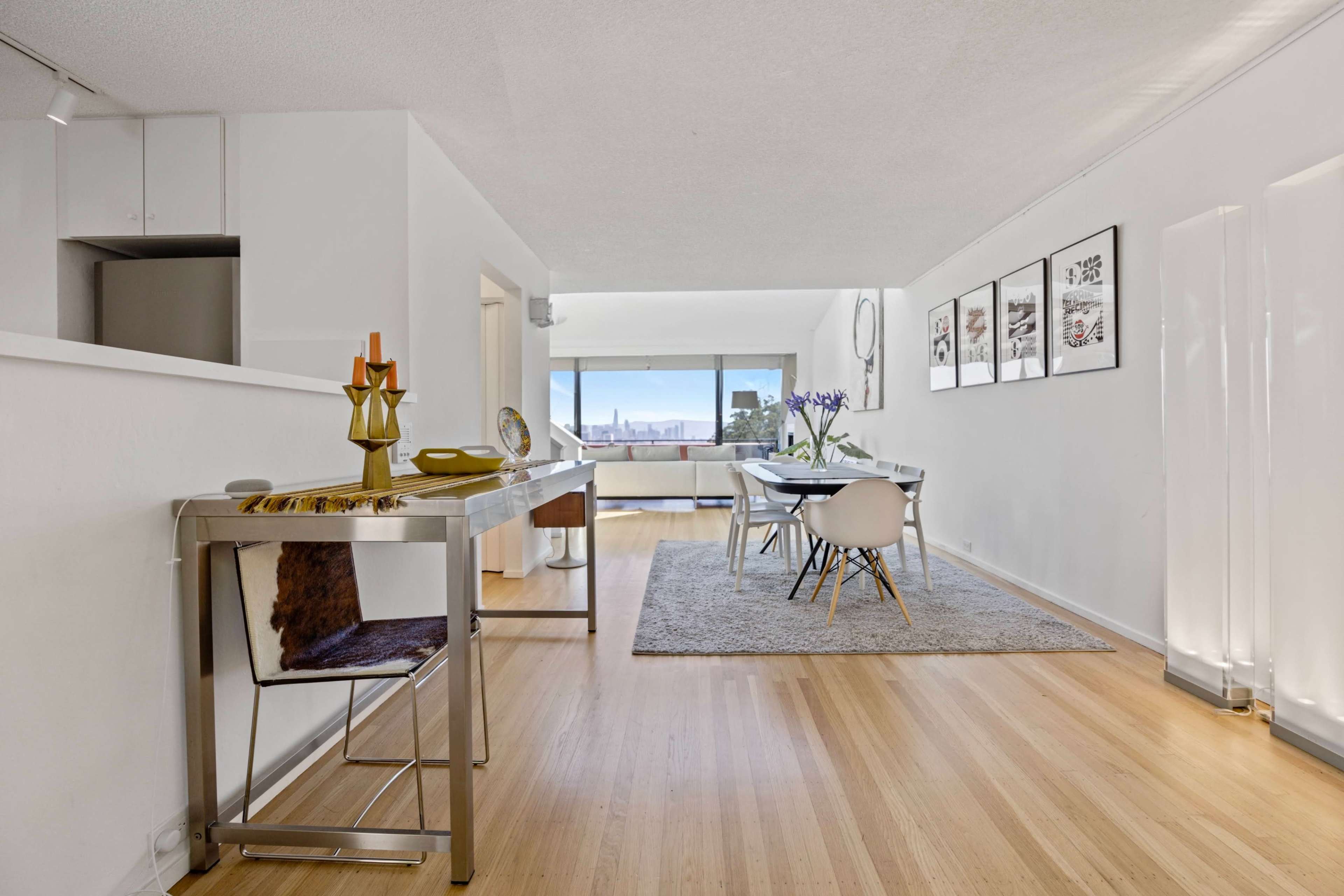 The image shows a bright, modern interior with a living space featuring a dining table, chairs, and a decorative console table.