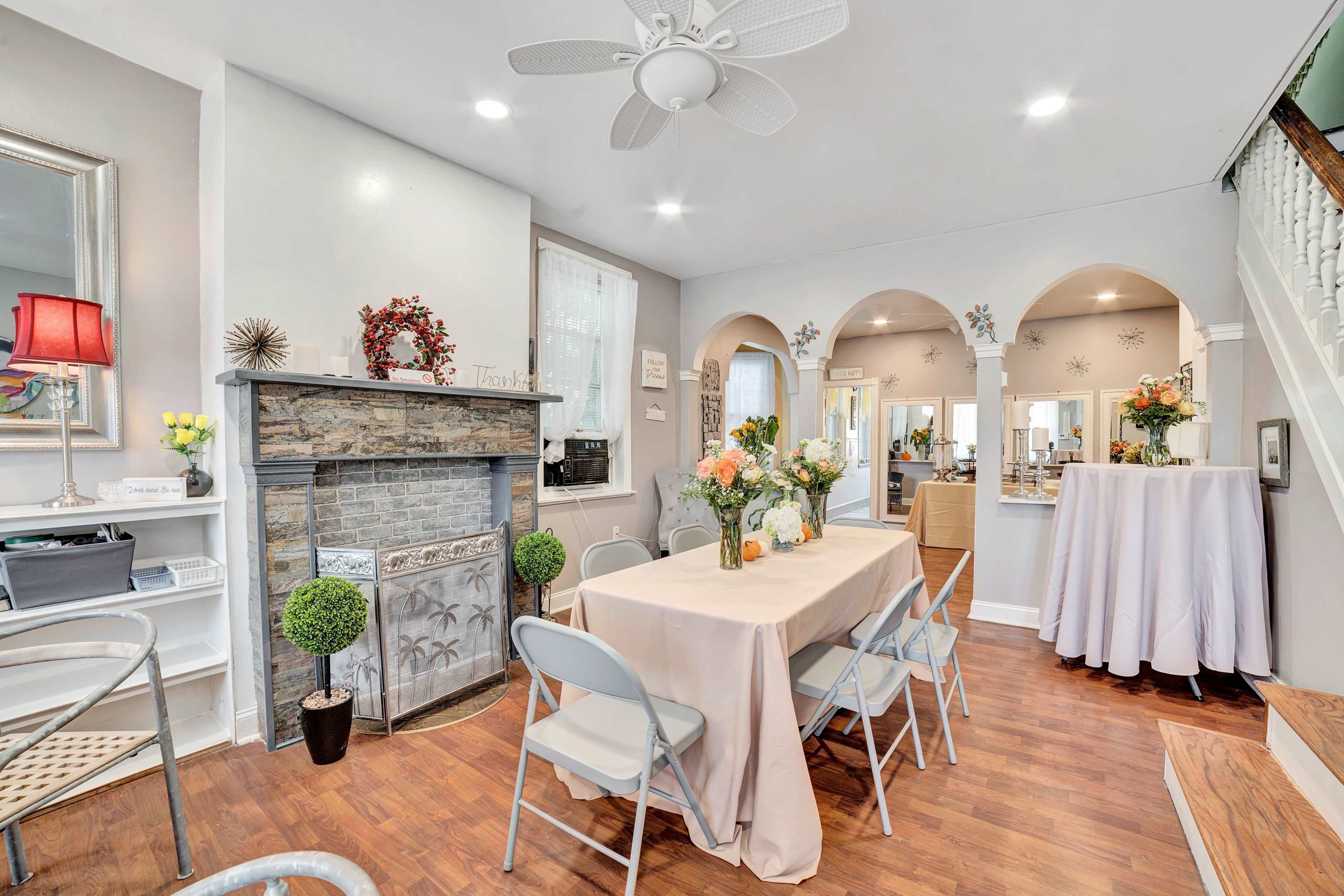 The image shows a cozy room with a dining table set for several people, a decorative fireplace, and a staircase leading to another area.