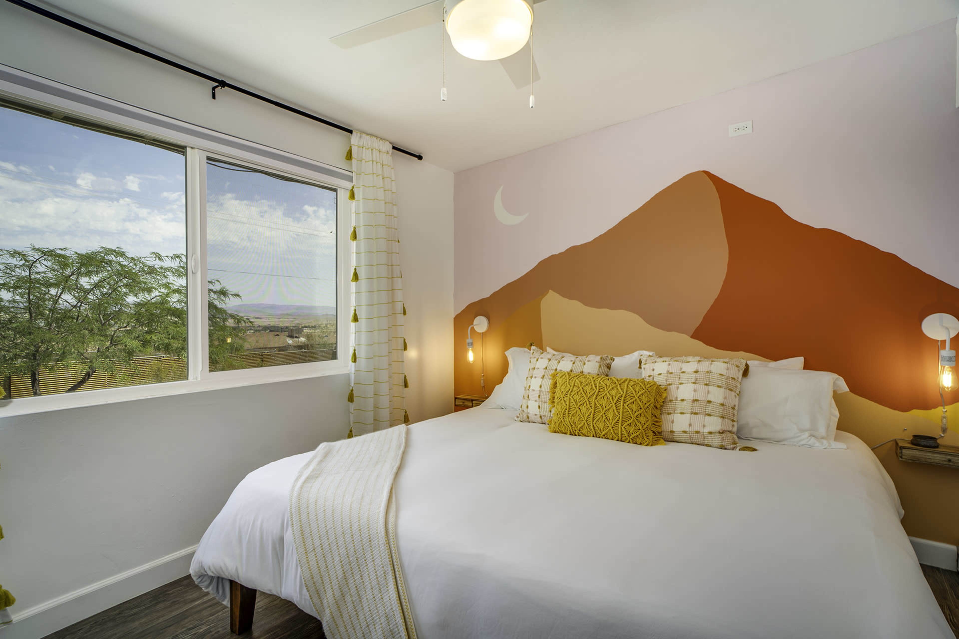 The image shows a well-lit bedroom with a large bed featuring decorative pillows and a mural of a mountain range on the wall.