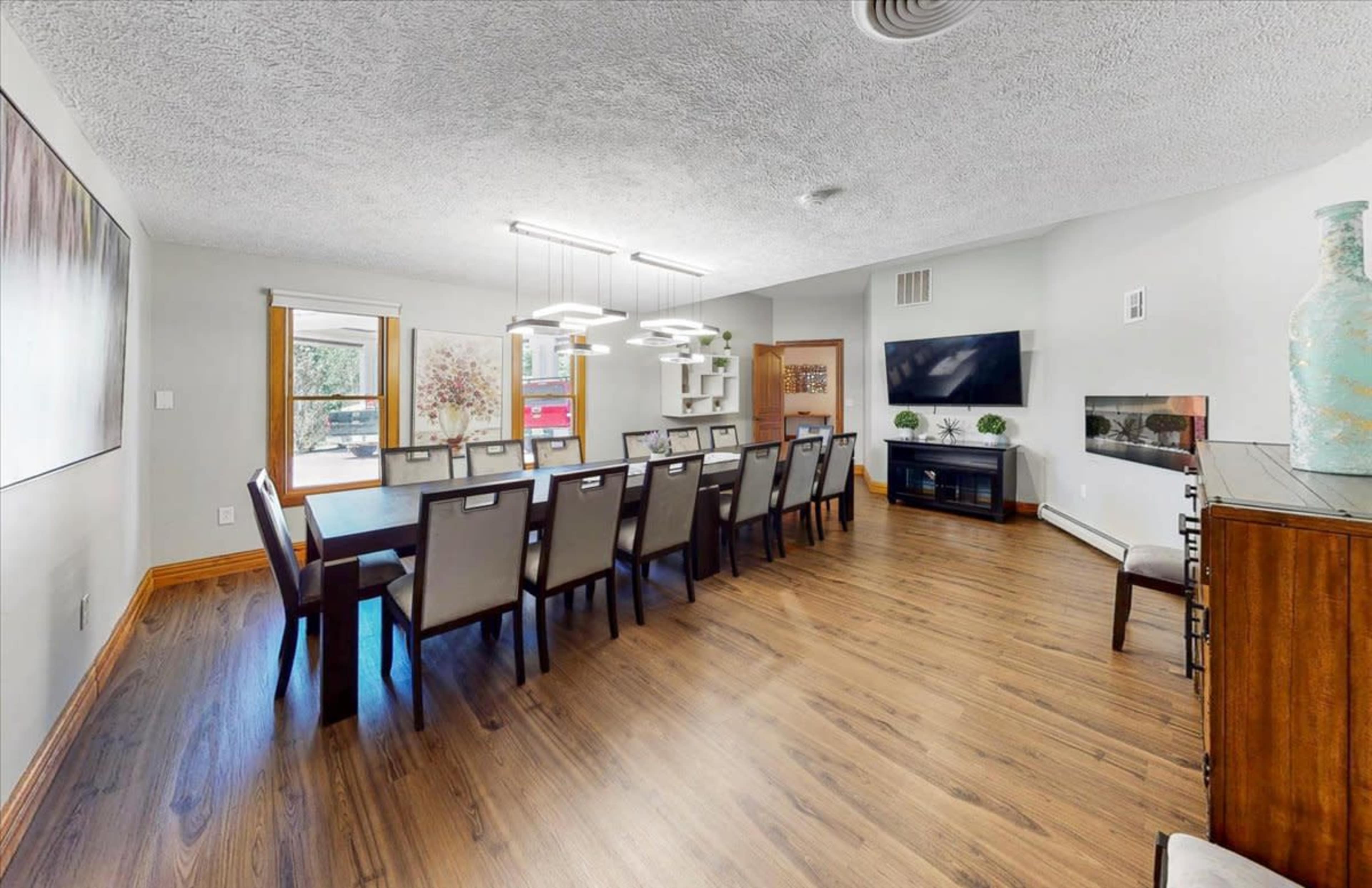 A long dining table with ten chairs is set in a spacious room featuring a TV, shelving unit, and large windows.