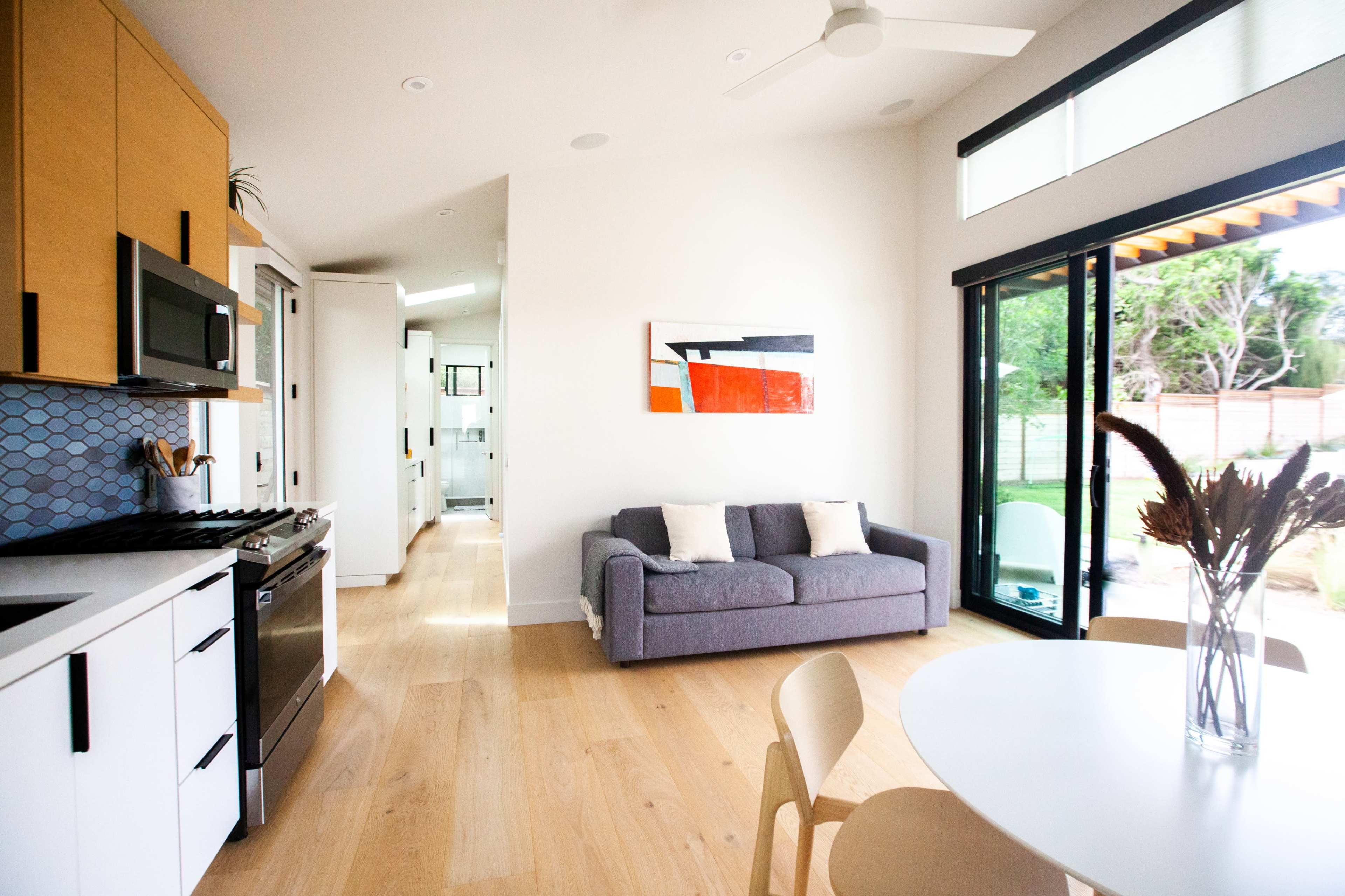 The image shows a modern kitchen and dining area with light wood flooring, a gray sofa, and large windows opening to an outdoor space.