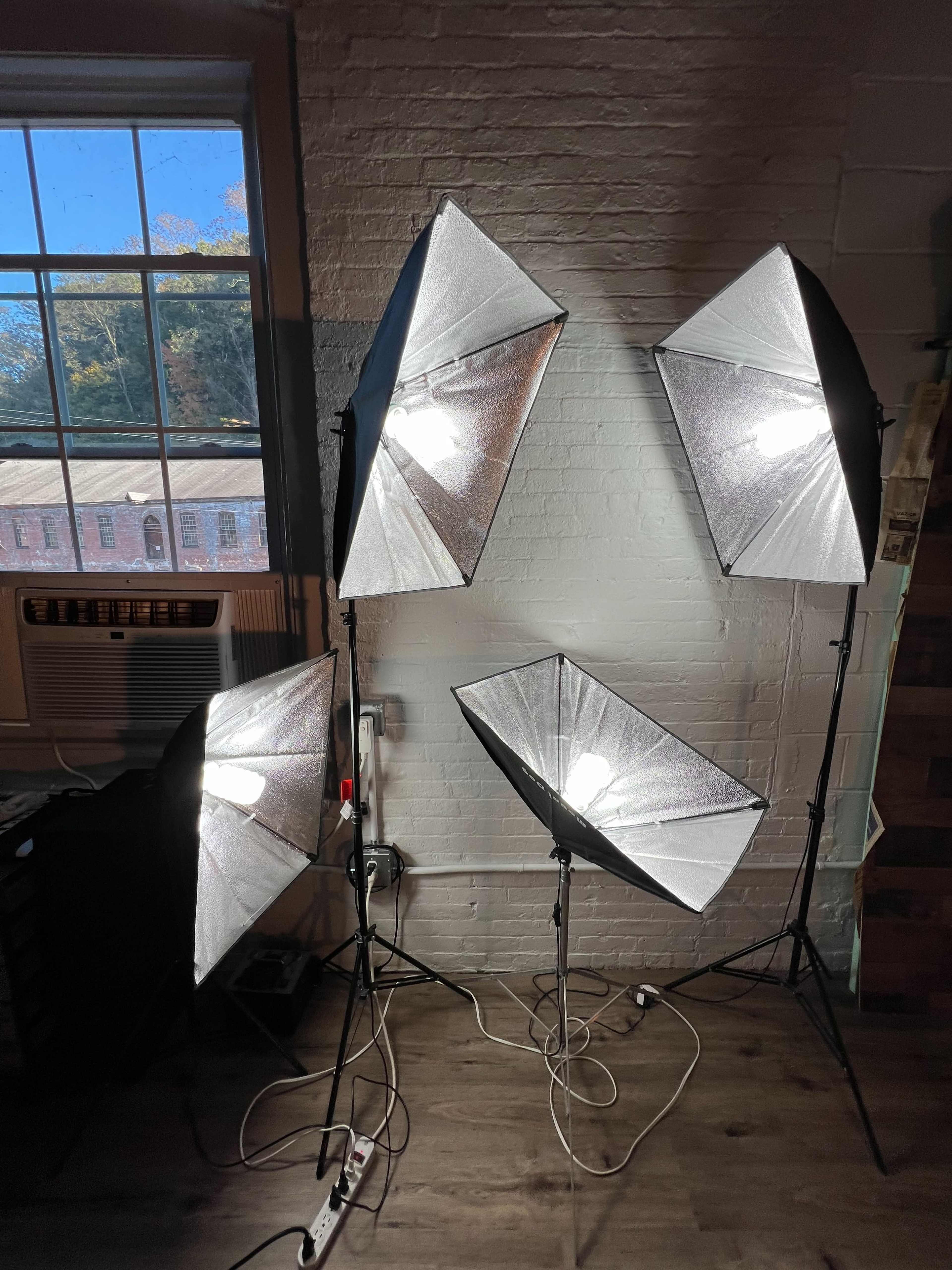 Four softbox lighting setups arranged in a well-lit room with a window and an air conditioning unit in the background.
