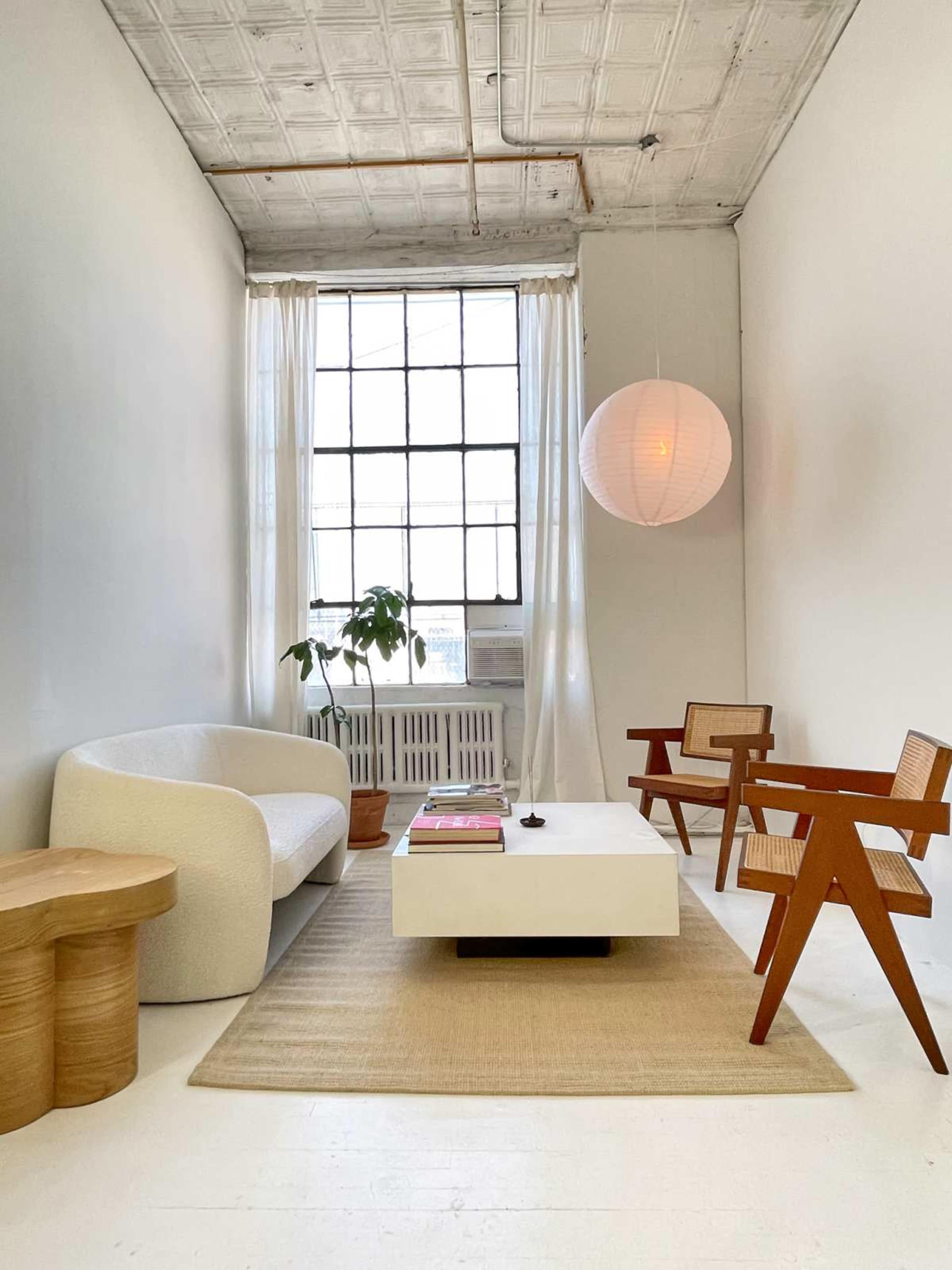 The image shows a minimalist living room featuring a white curved sofa, two wooden chairs, a low coffee table, and a large window with sheer curtains.