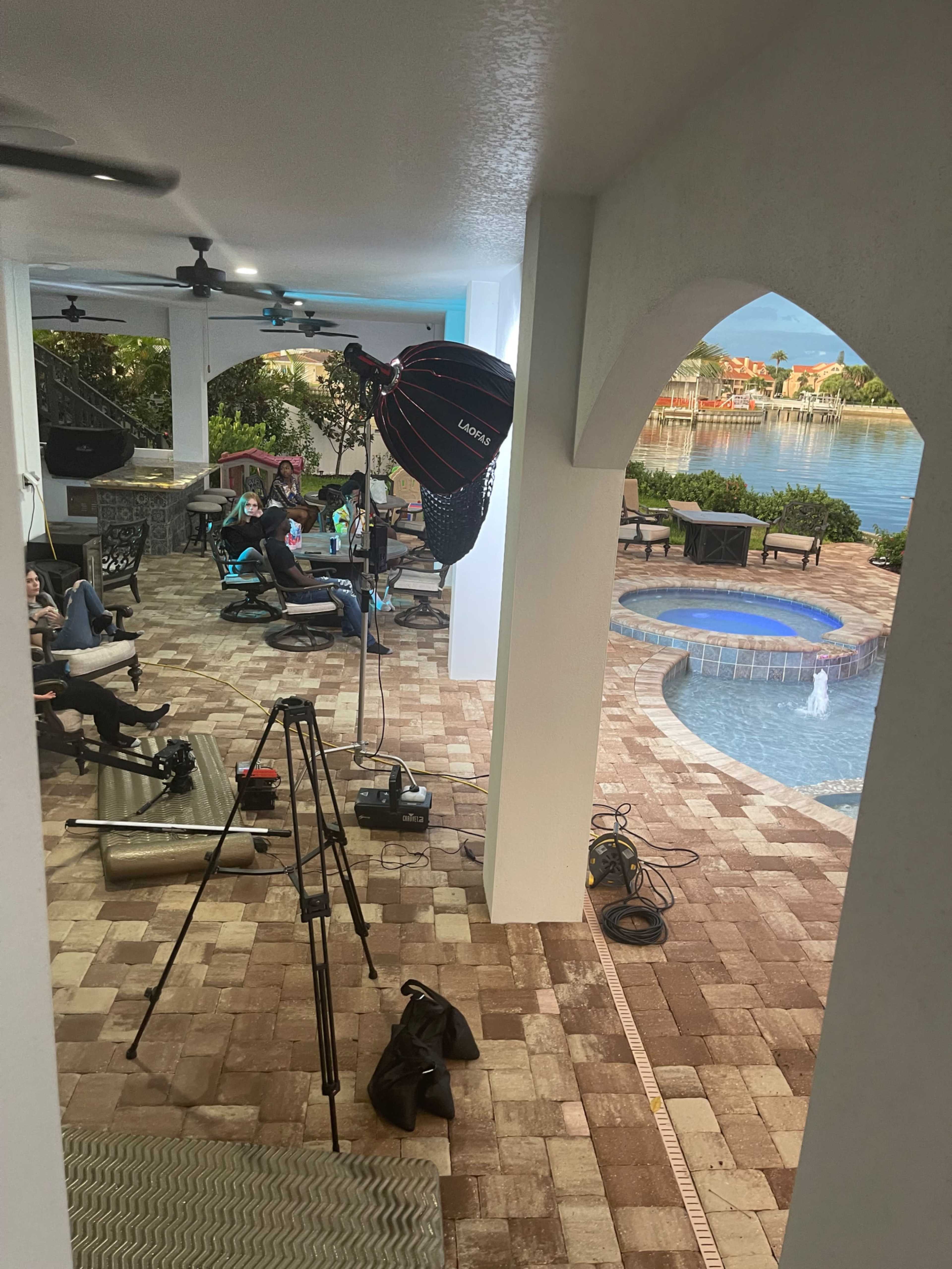 The image shows a poolside area with lounging chairs, photography equipment, and a view of the water and houses in the background.
