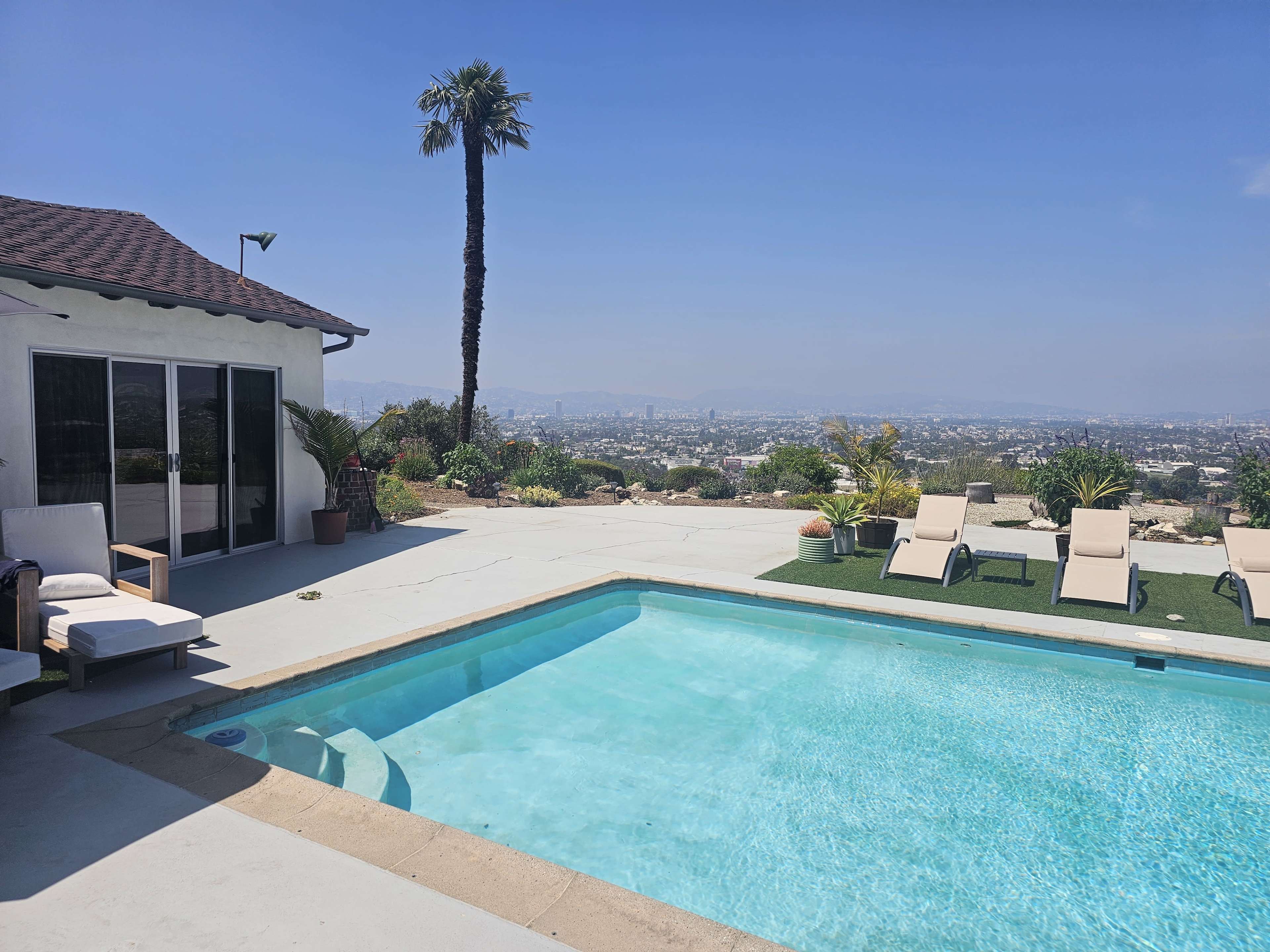 A swimming pool sits in the foreground of a hillside home, with lounge chairs arranged around it and a view of the city in the background.