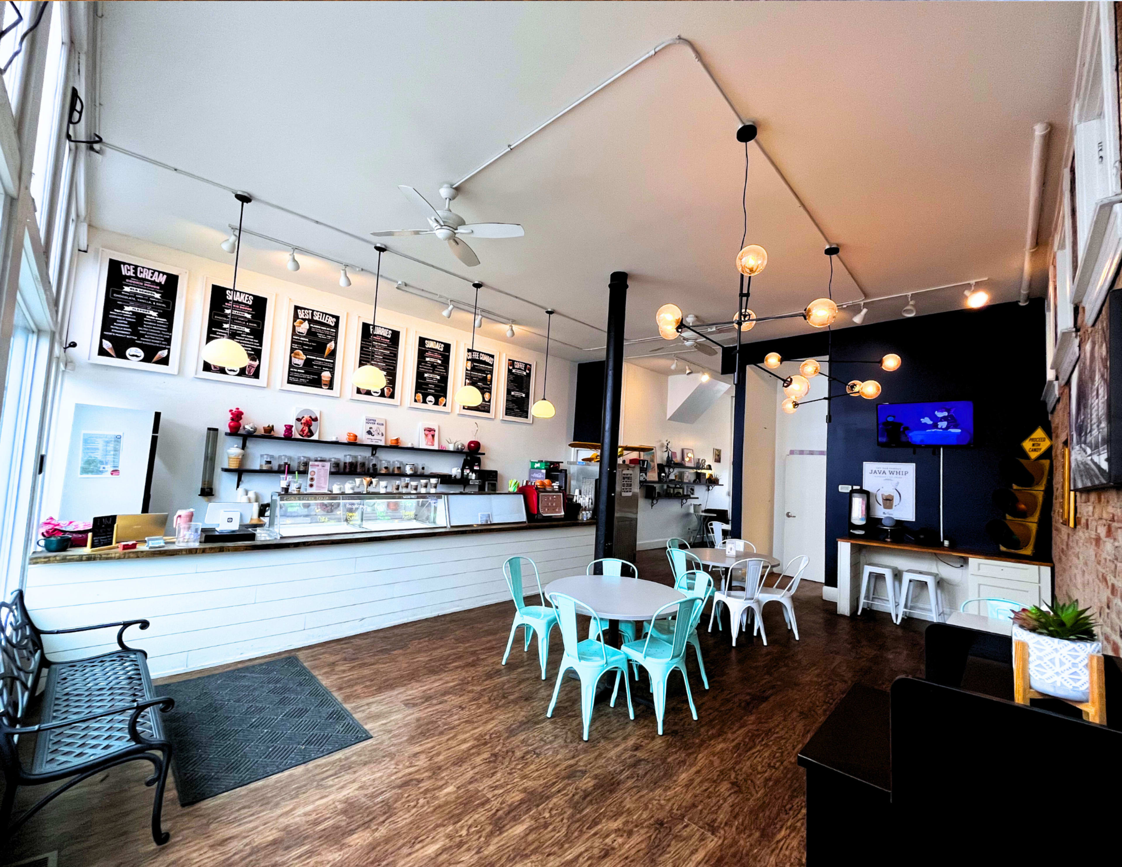 The image shows a modern ice cream cafe with light wooden floors, turquoise chairs and tables, a service counter displaying a variety of ice cream flavors, and bright lighting fixtures.