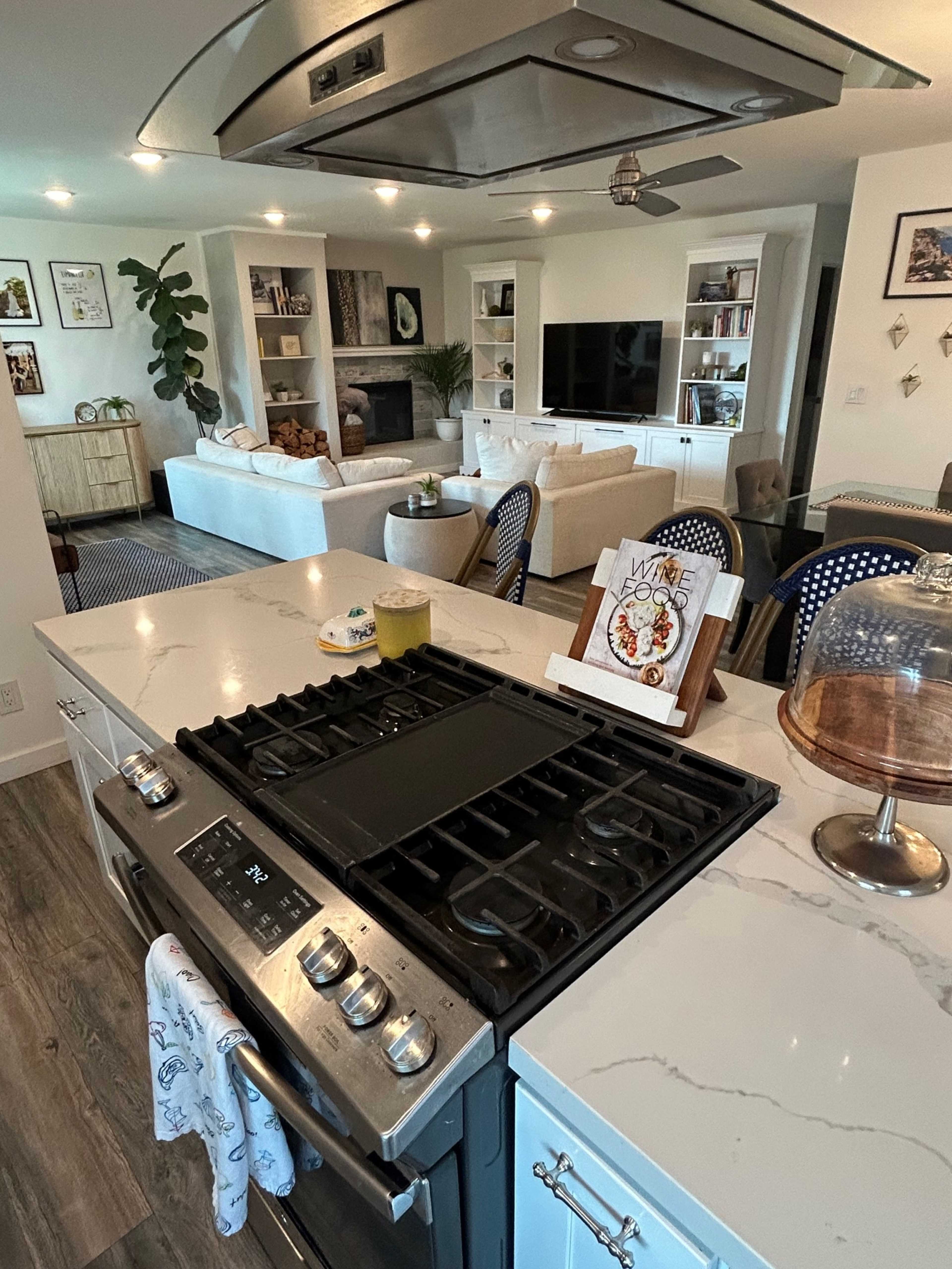 The image shows a modern kitchen with a gas stove and an island that looks into a spacious living room furnished with a sofa and a coffee table.