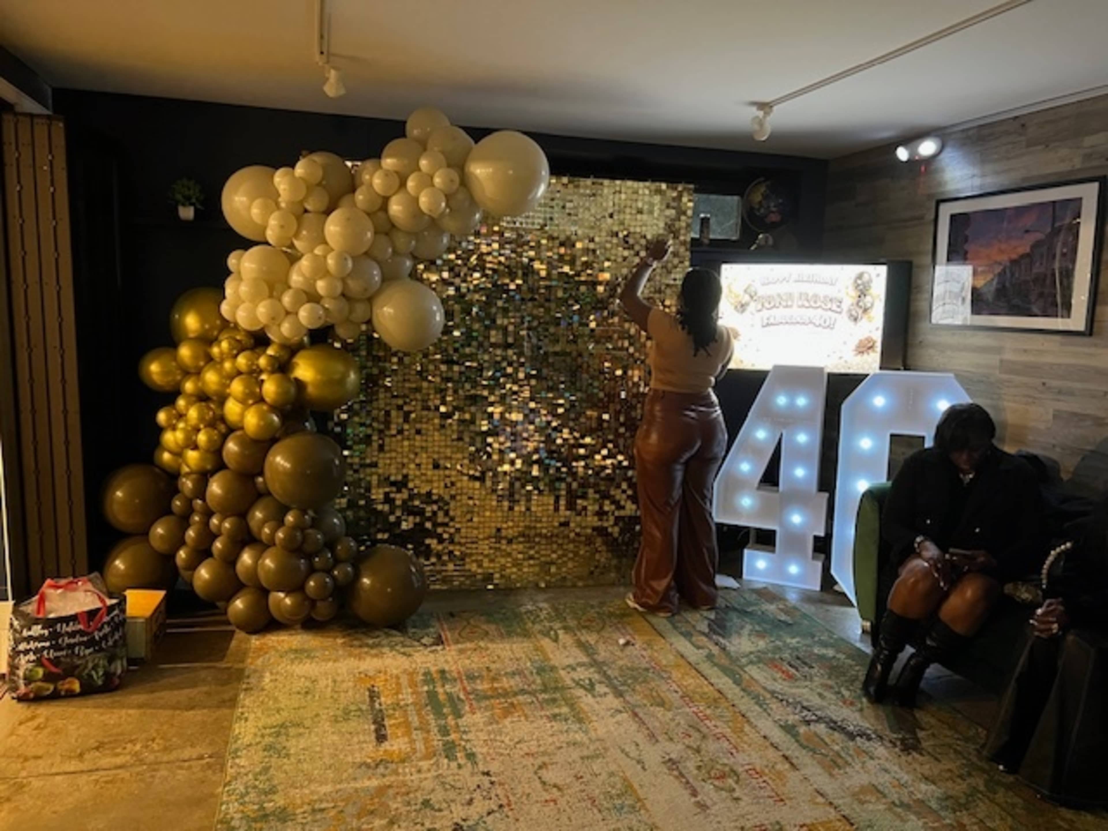 A person arranges balloons near a golden sequin backdrop at a decorated event space with illuminated numbers displaying "40."