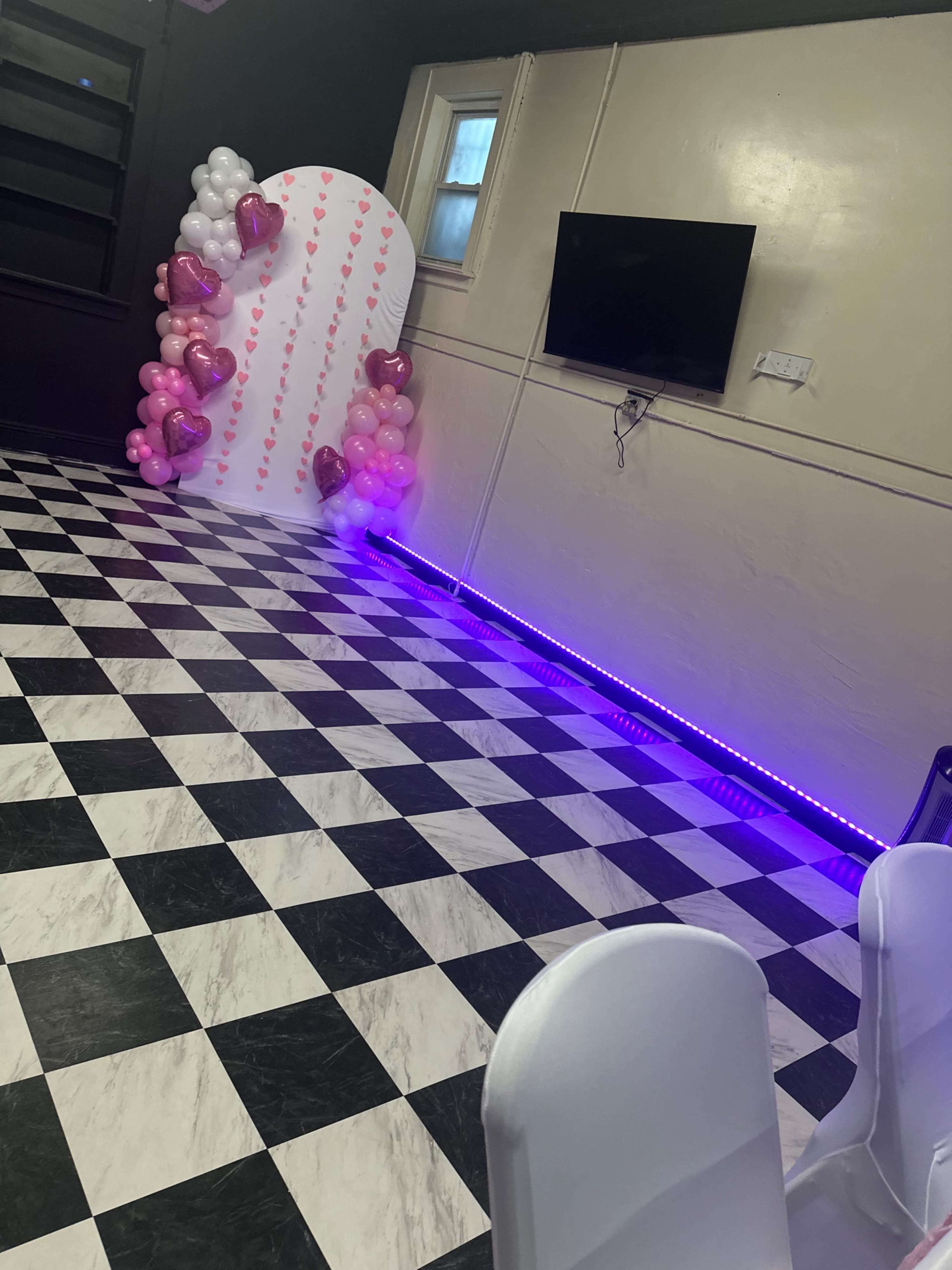 The image shows a decorated room with a black and white checkered floor, a large white backdrop decorated with pink and gold balloons, and purple LED lighting along the base.
