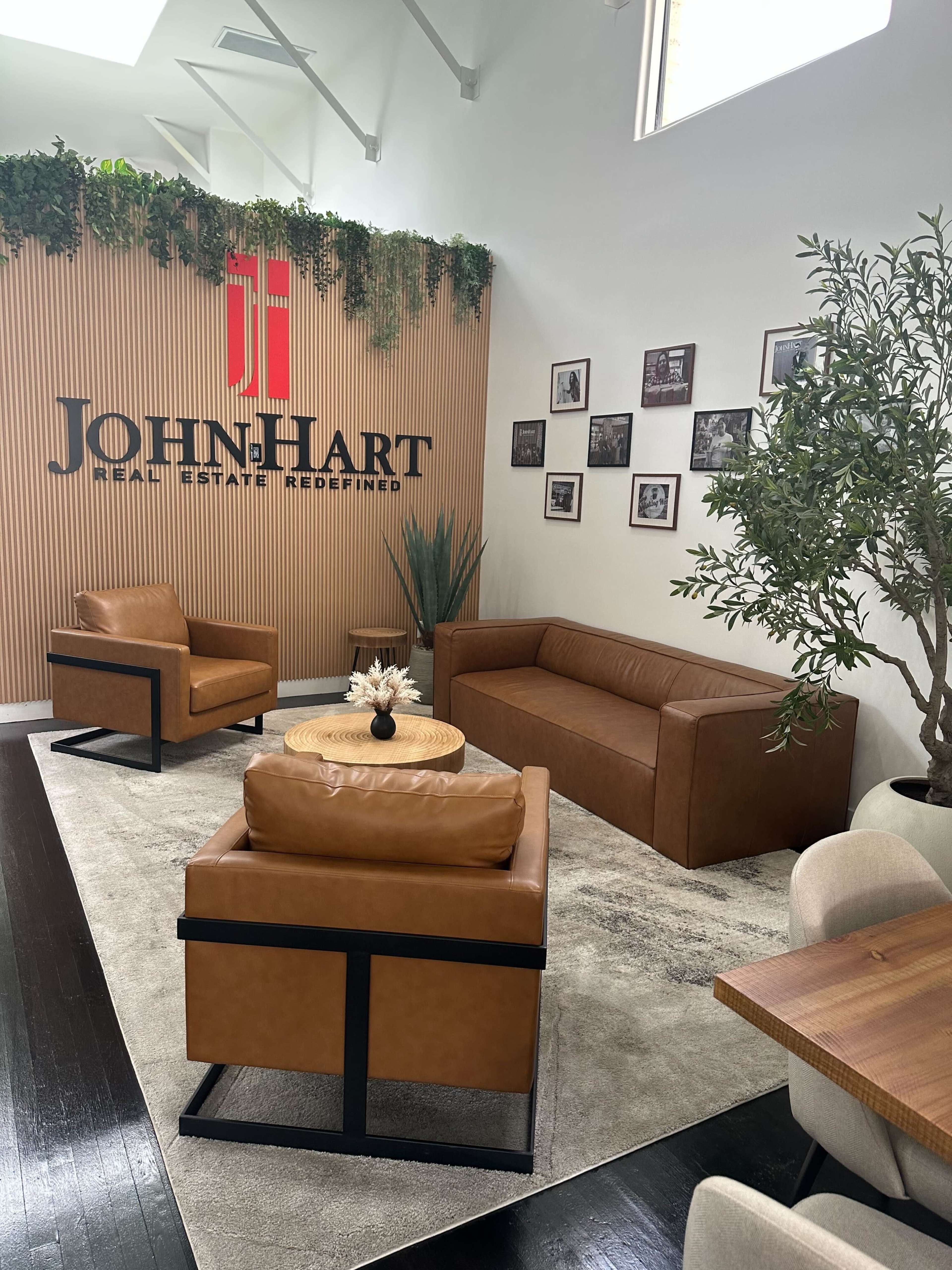 The image shows a modern real estate office lobby featuring brown leather seating, a wooden accent wall with the JohnHart logo, and framed photographs on the walls.