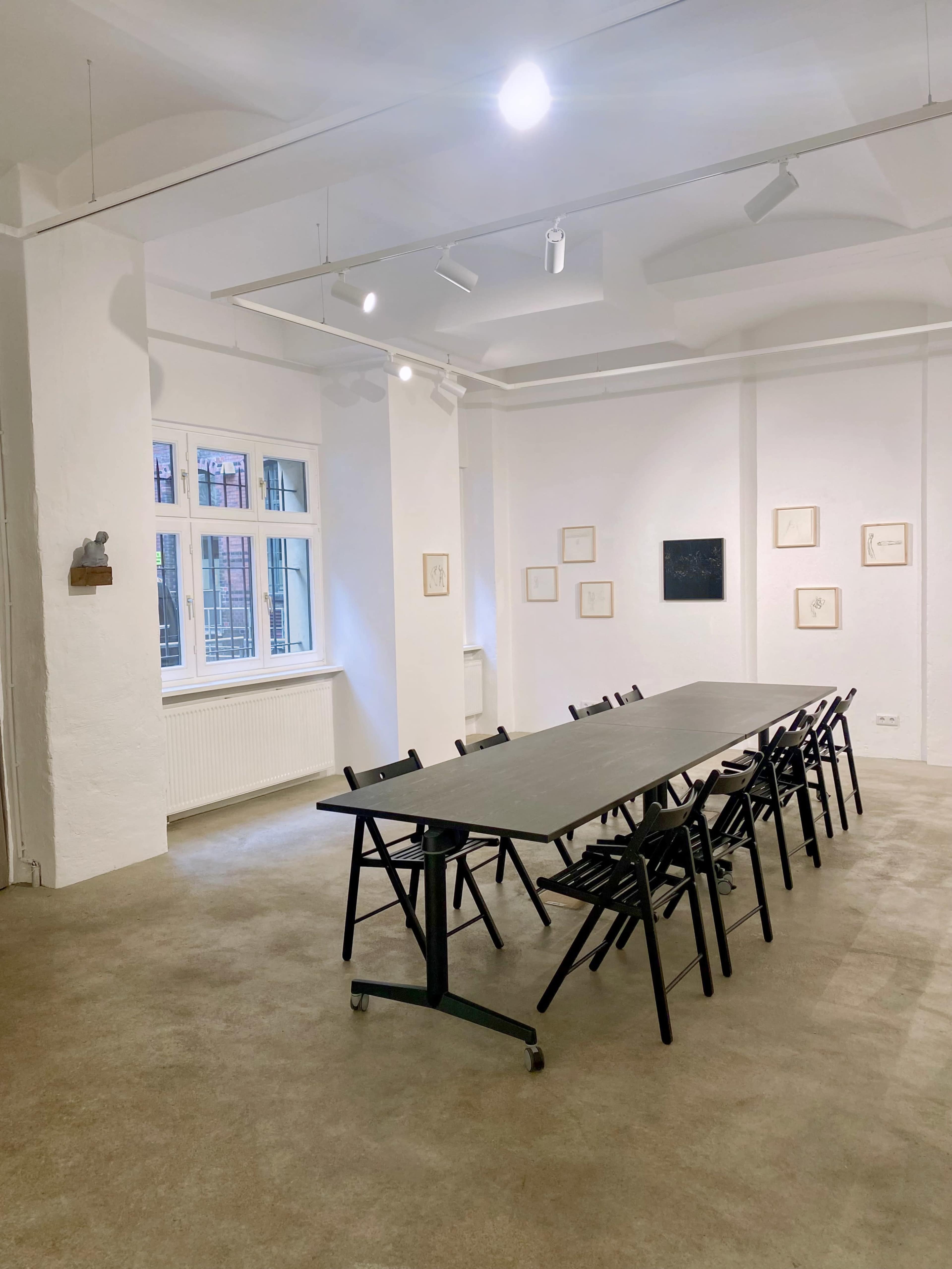 A long table surrounded by black chairs is set in a well-lit, minimalist room with white walls and large windows.
