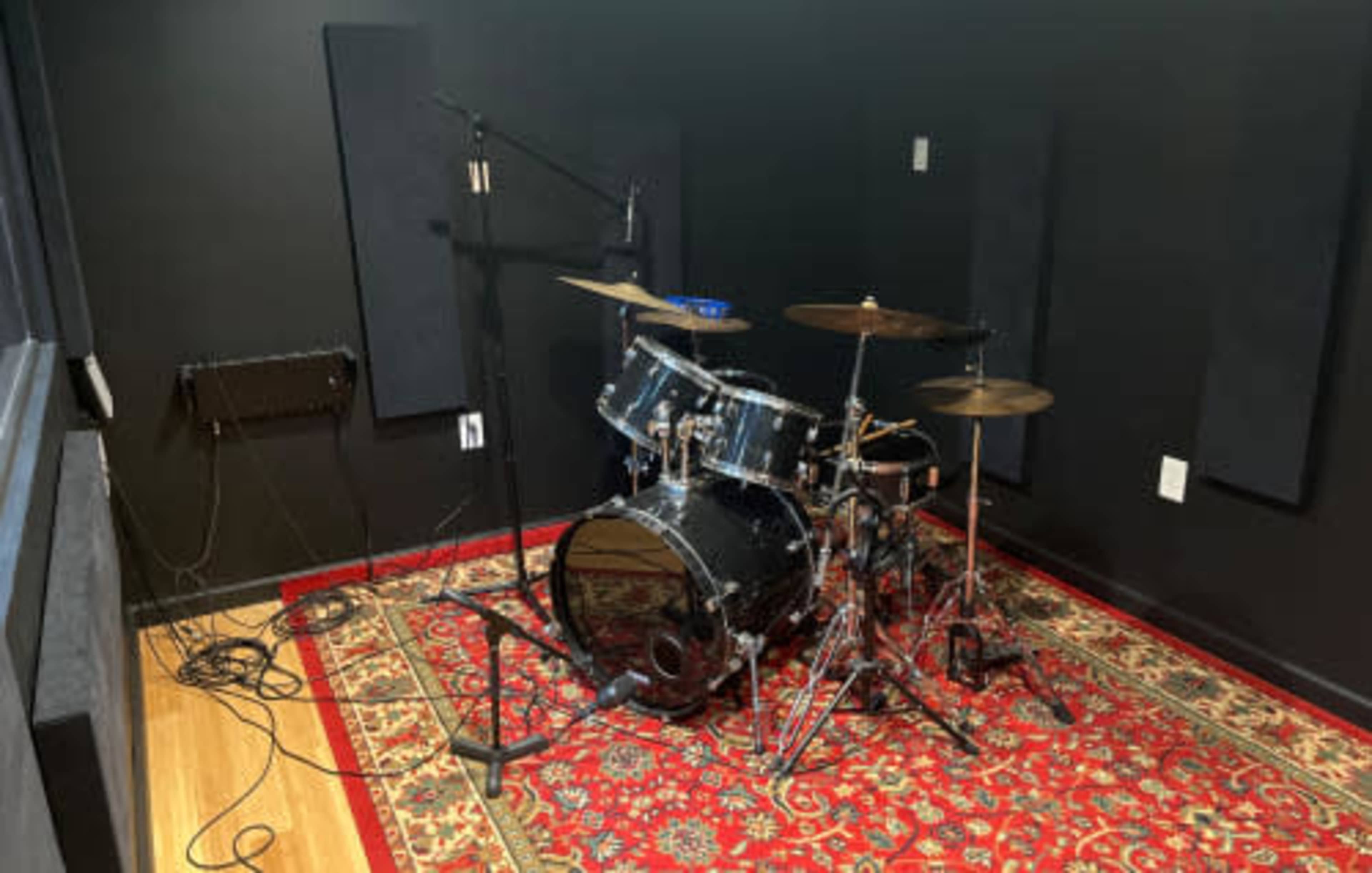 A drum kit is set up on a patterned rug in a soundproofed rehearsal room with black walls and acoustic panels.