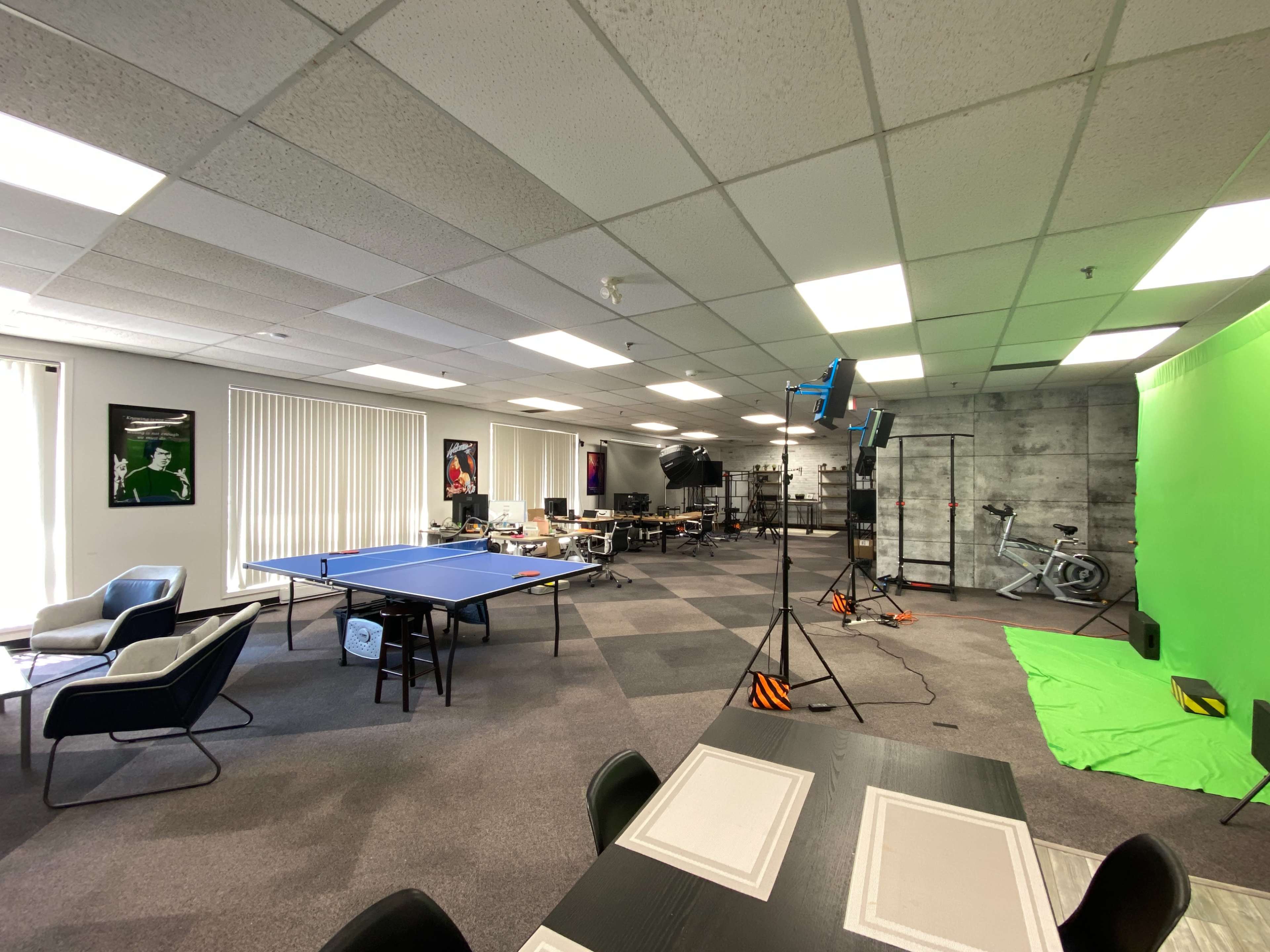 A spacious office area featuring a ping pong table, a green screen setup, and various workstations.