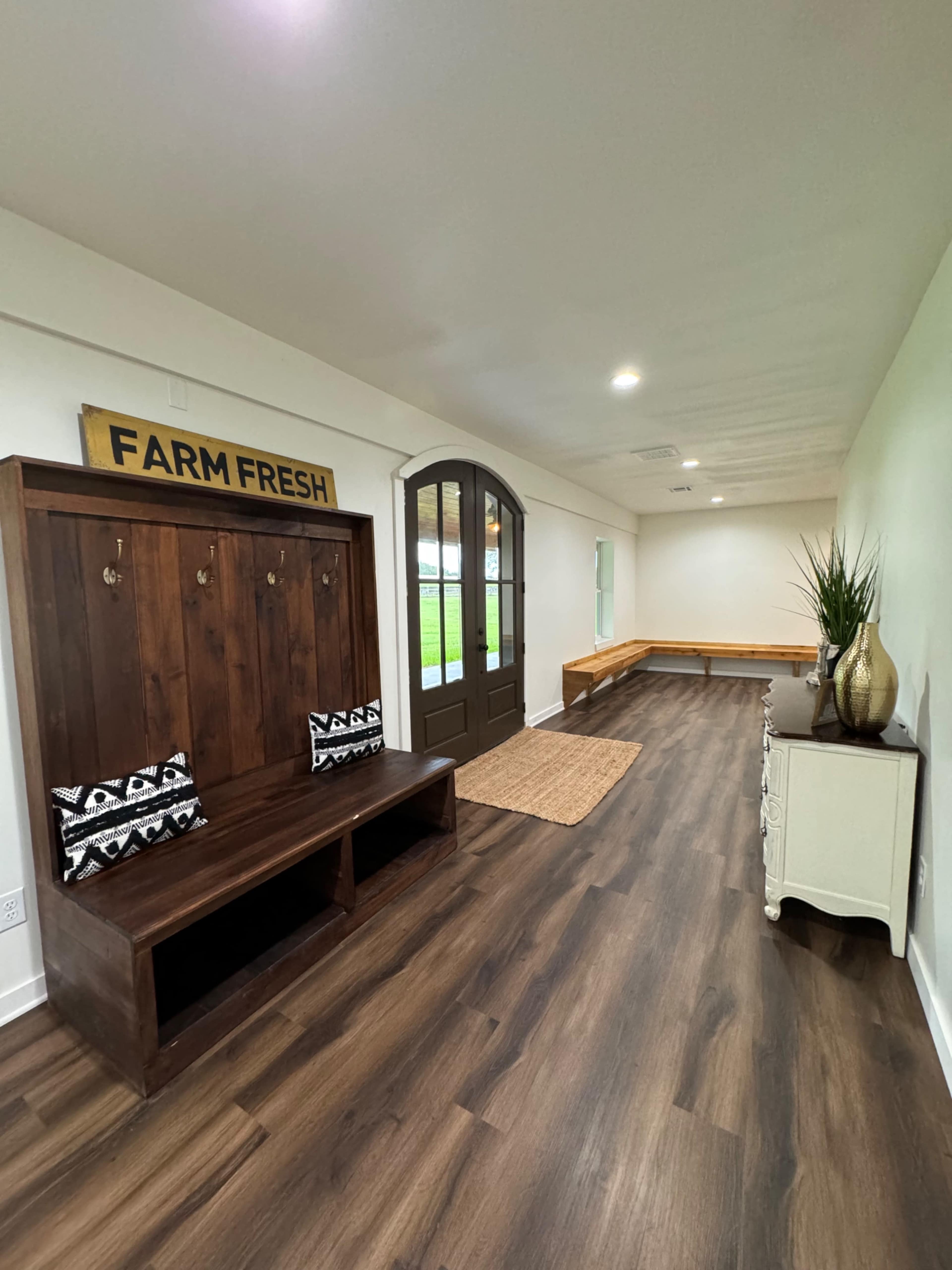A hallway with a wooden bench and coat hooks on one side, a set of double doors at the end, and a small table with decorative items on the opposite side.