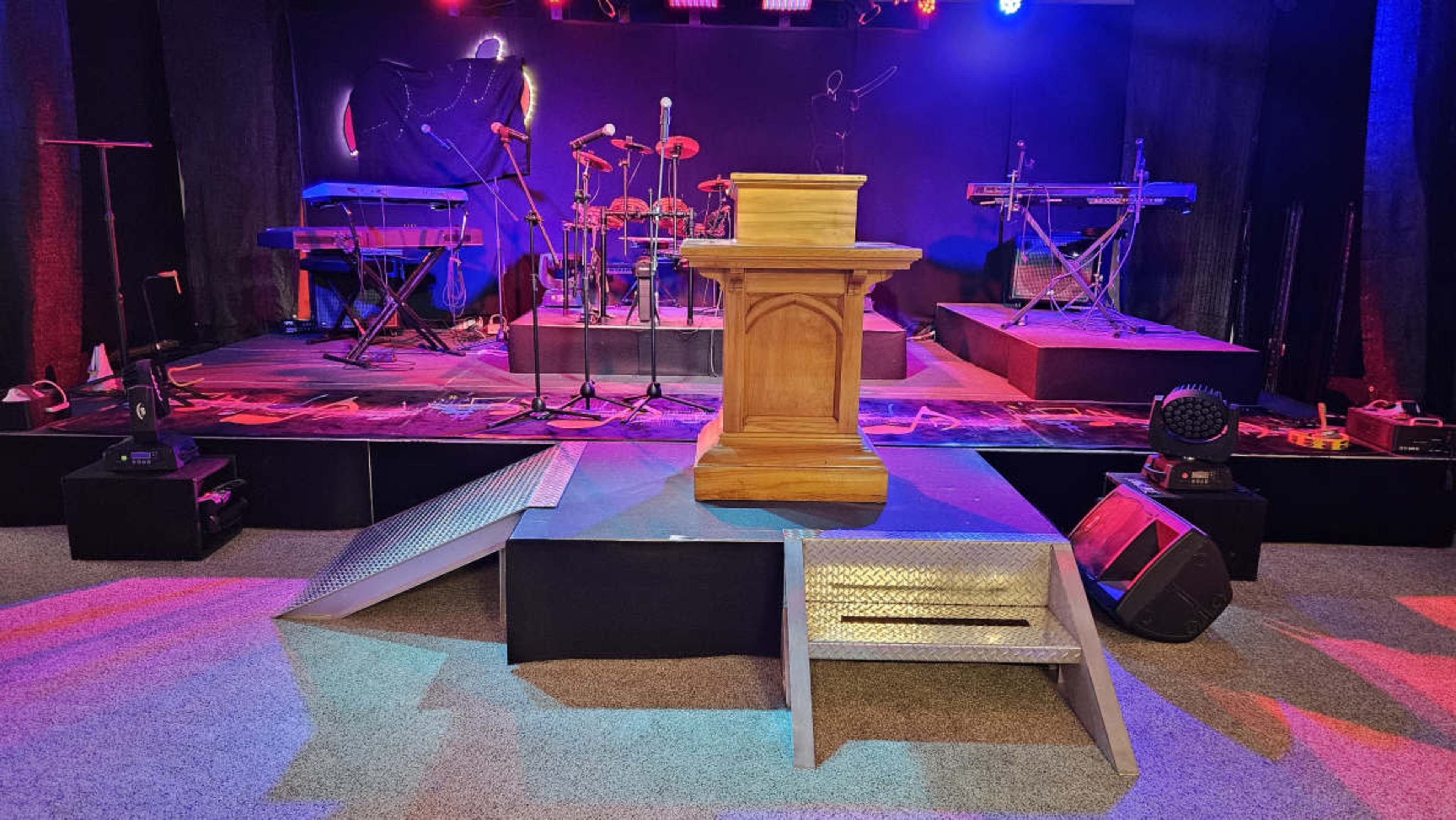 A stage setup with a wooden podium at the center, flanked by musical instruments and equipment under colorful stage lighting.