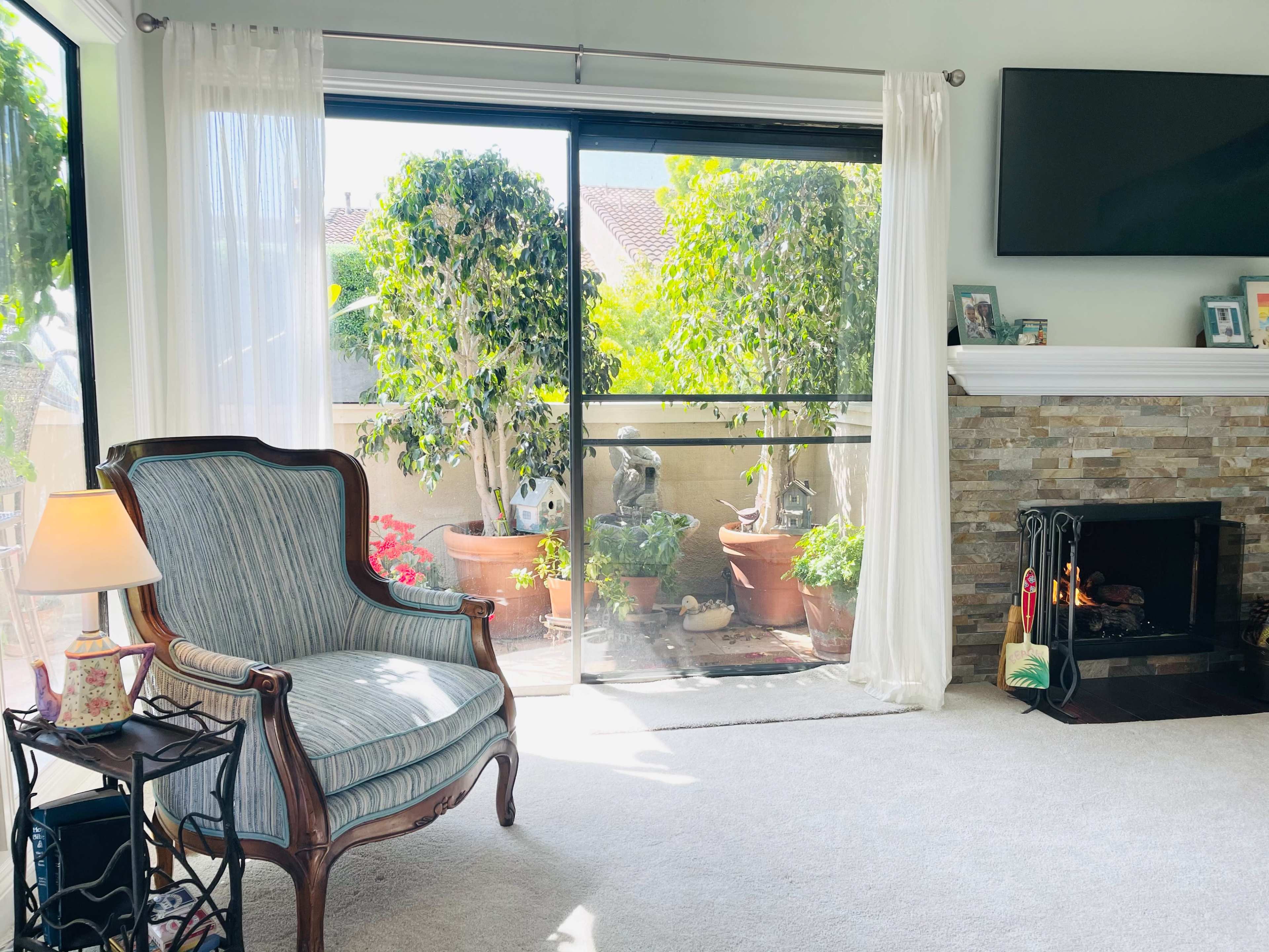 A cozy living room features a striped armchair beside a small lamp, with large windows revealing a balcony garden filled with potted plants.
