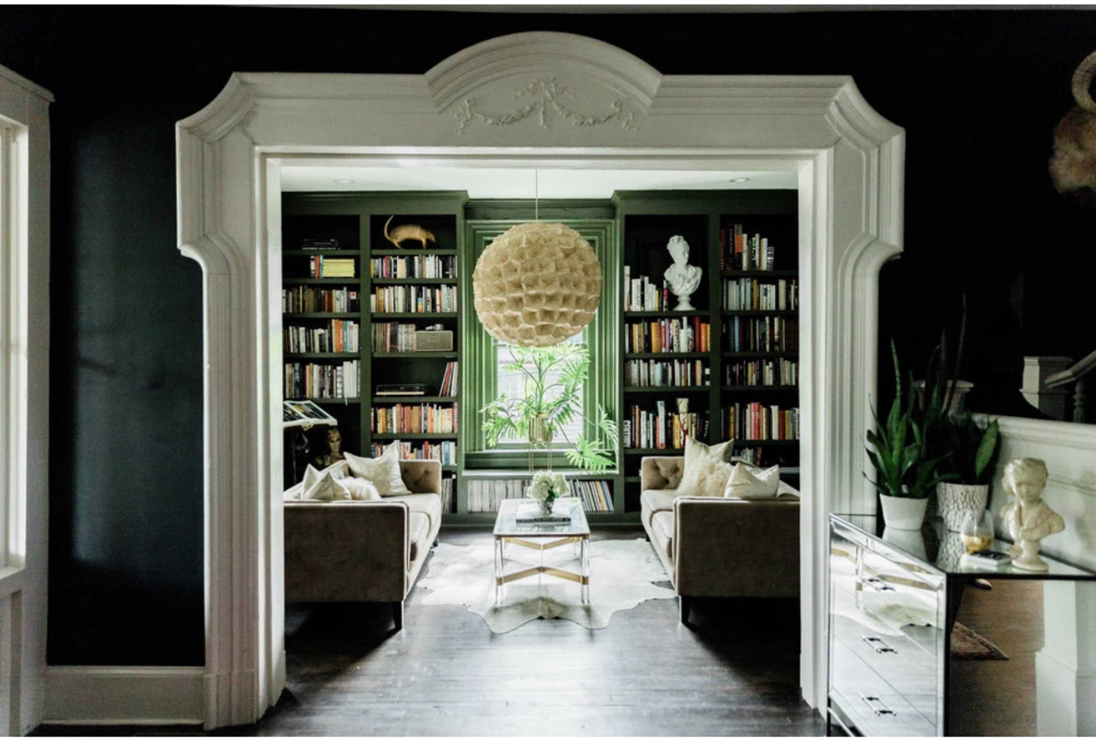 A modern living room features a large bookshelf filled with books, two sofas with decorative pillows, and a pendant light hanging from a detailed archway.