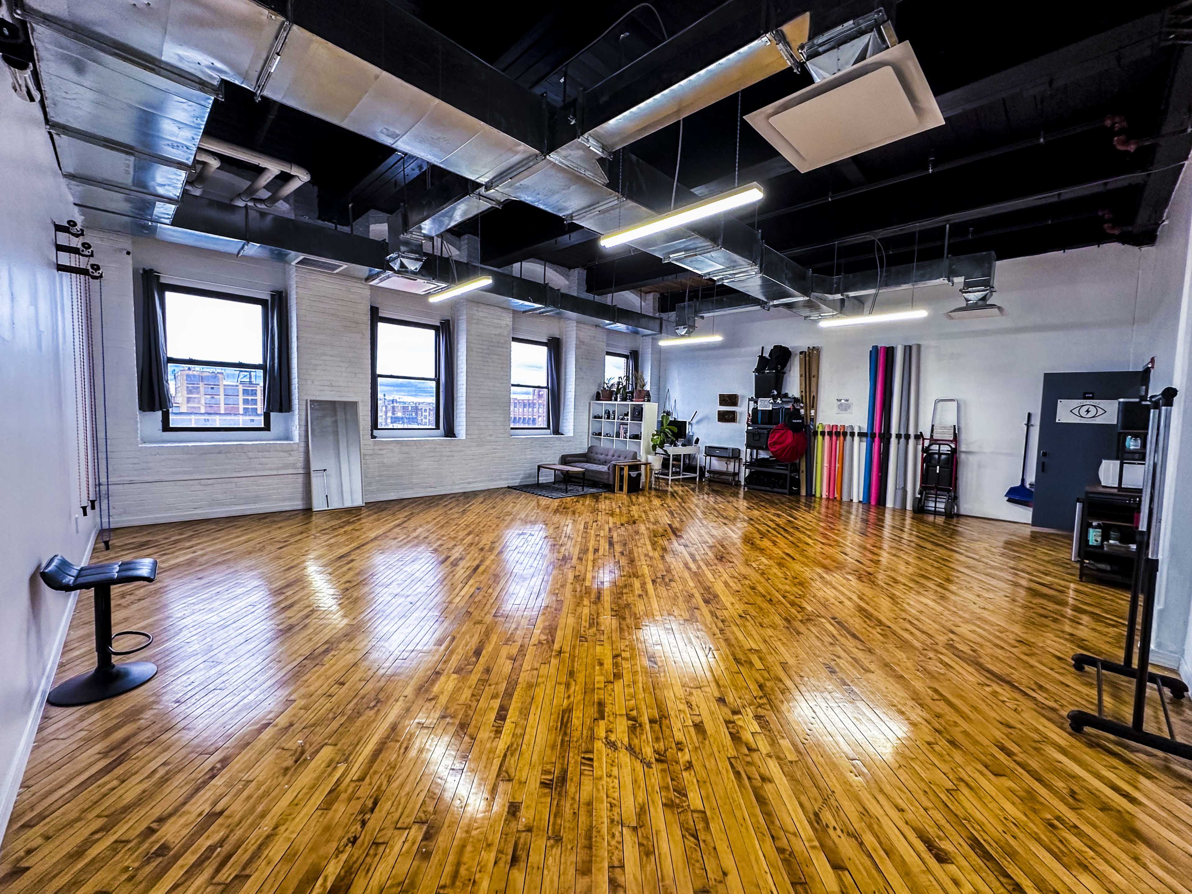 A spacious, empty studio with polished wooden floors, large windows, and various equipment along the walls.