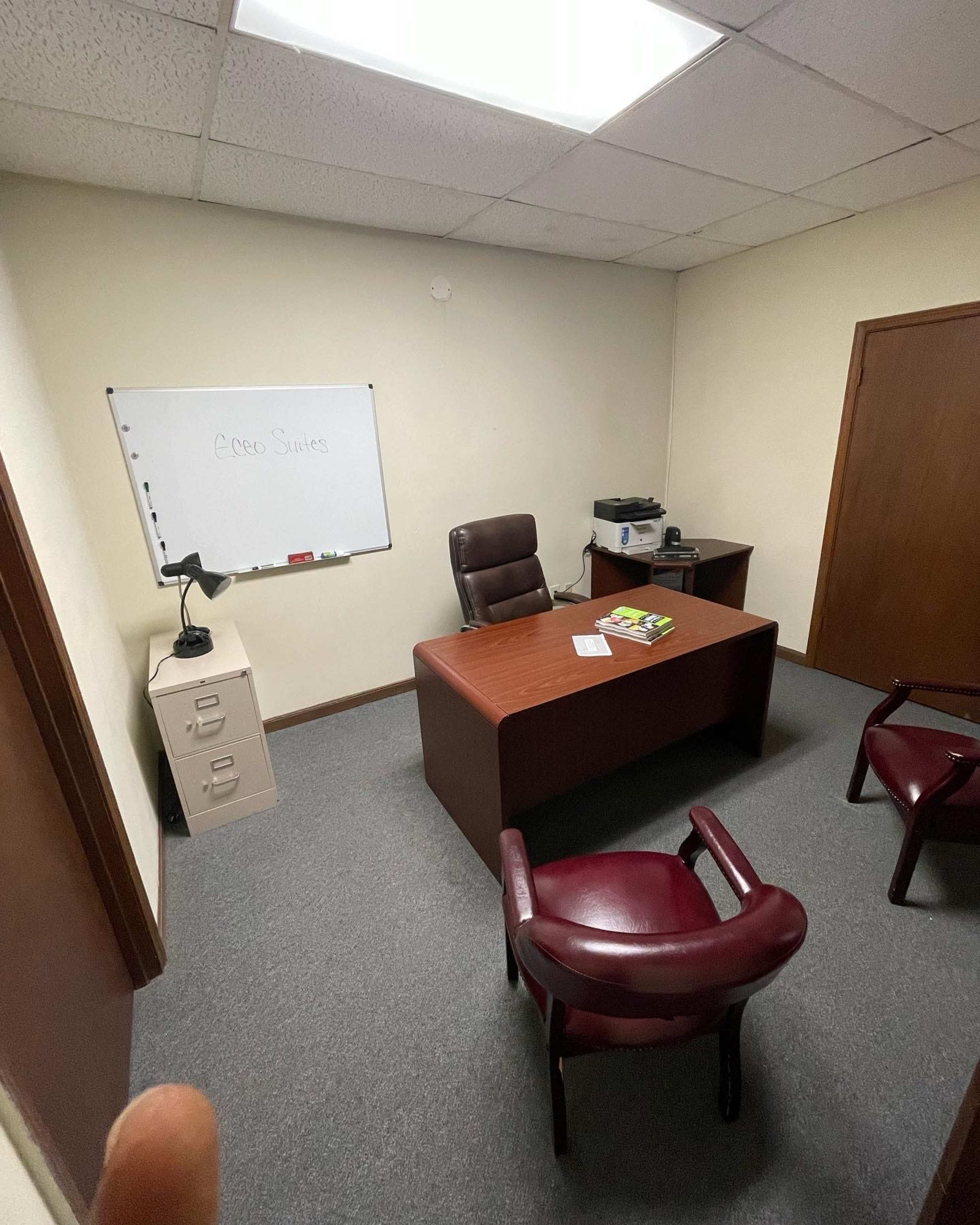 The image shows a small office with a wooden desk, two red chairs, a whiteboard, and a printer in the corner.