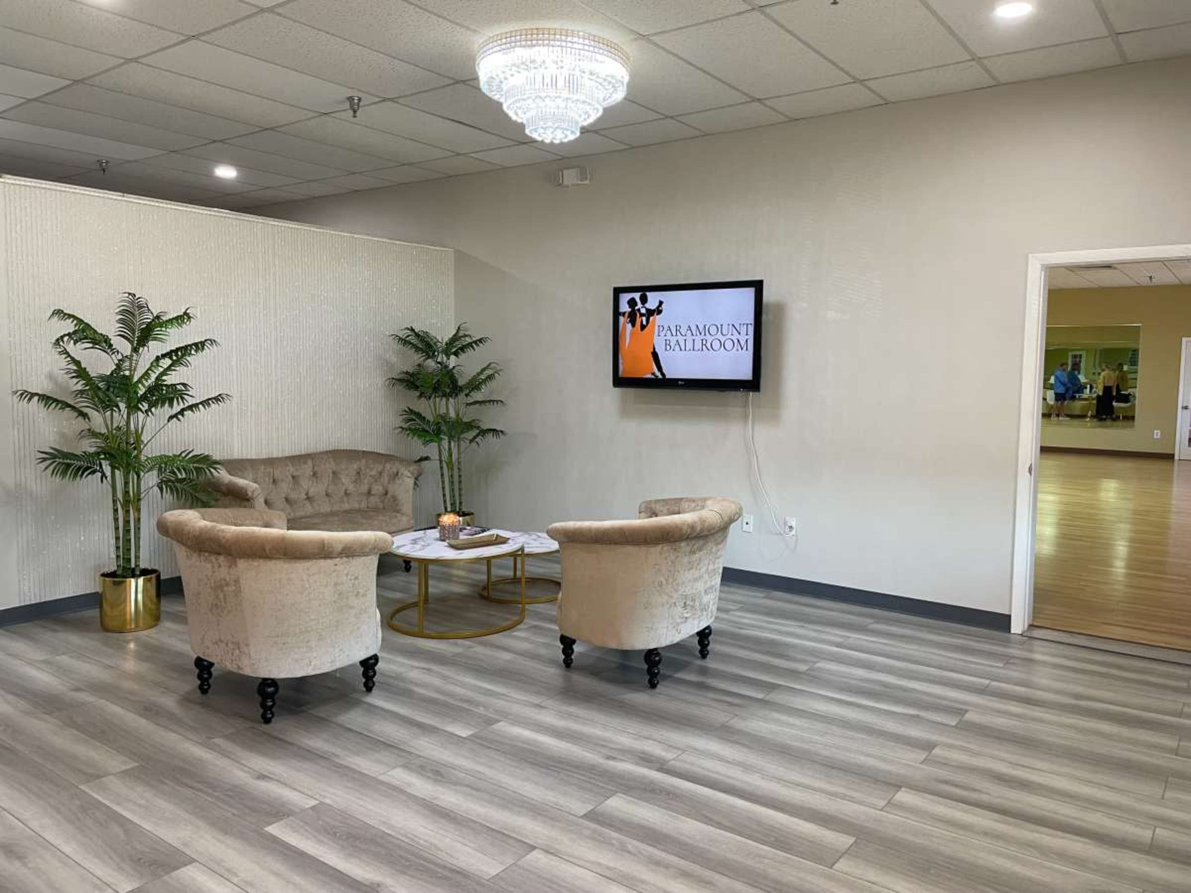 A waiting area with two upholstered chairs facing a coffee table, a television displaying "PARAMOUNT BALLROOM," and potted plants against a light-colored wall.