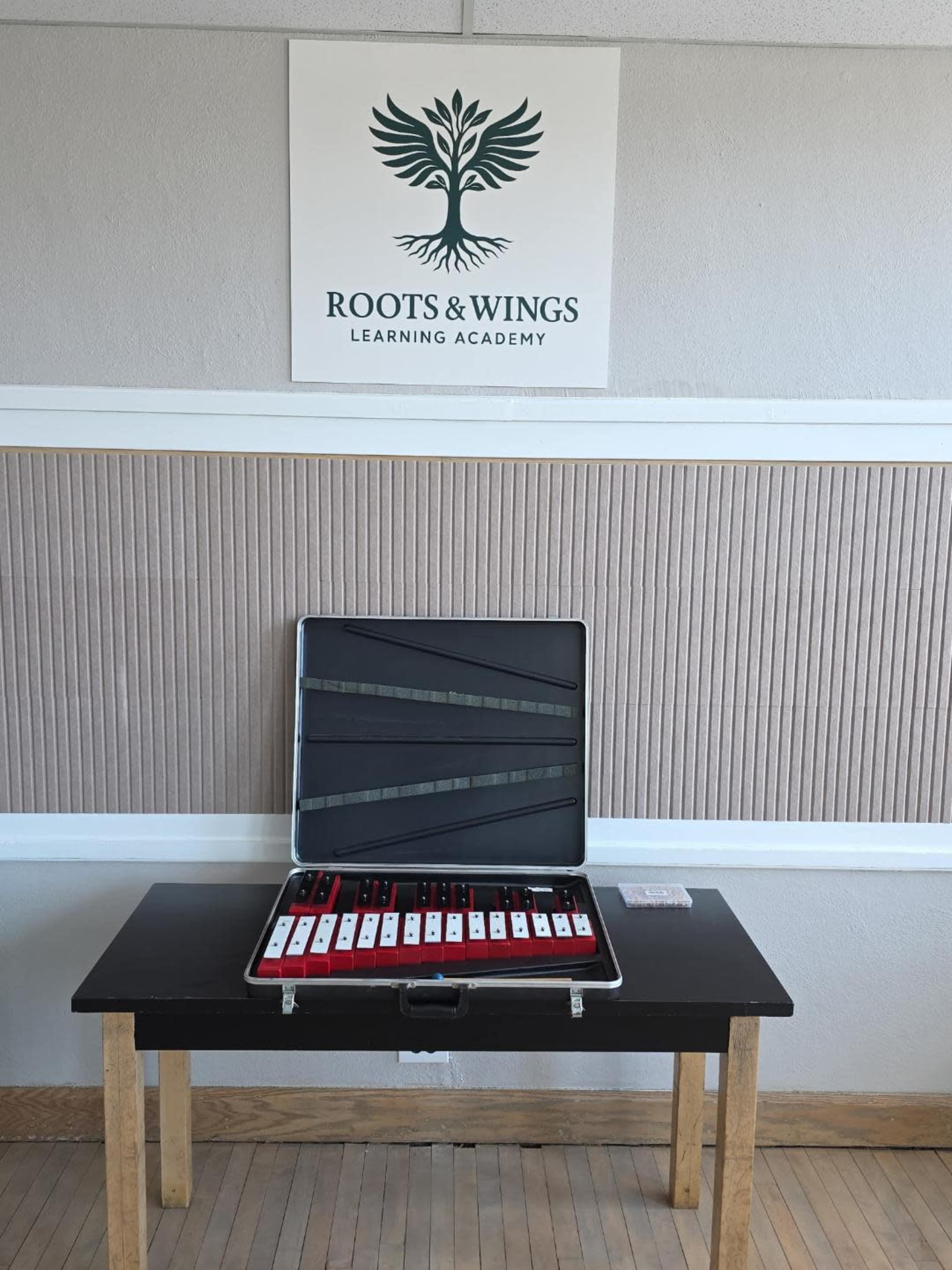A neatly arranged table displays a case of educational materials in front of a wall with a sign that reads "Roots & Wings Learning Academy."