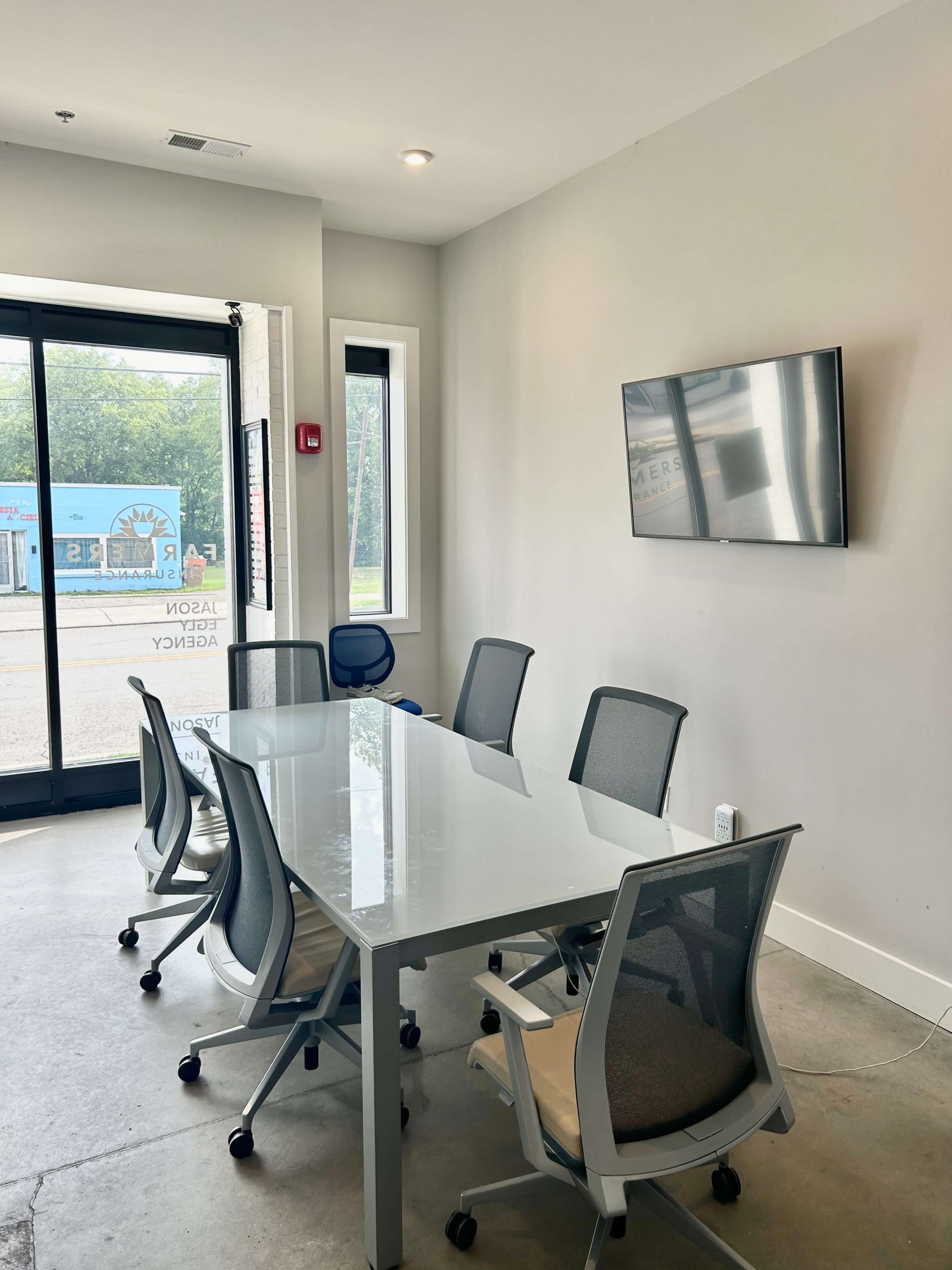 A modern conference room features a glass table surrounded by six ergonomic chairs and a wall-mounted television, with large windows providing natural light.