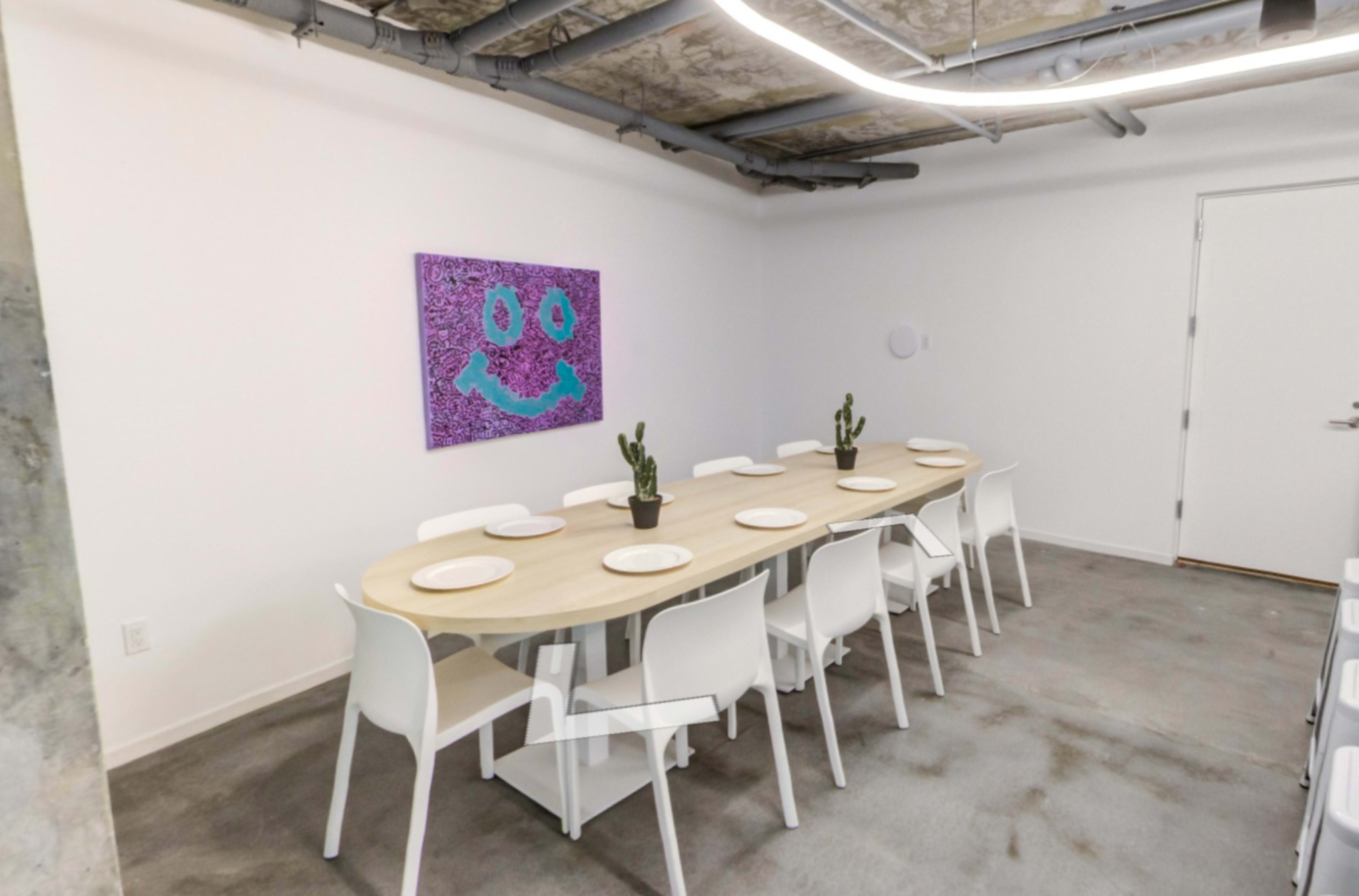 A modern dining area features a long, curved wooden table surrounded by white chairs, with a cactus centerpiece and a smiling face artwork on the wall.