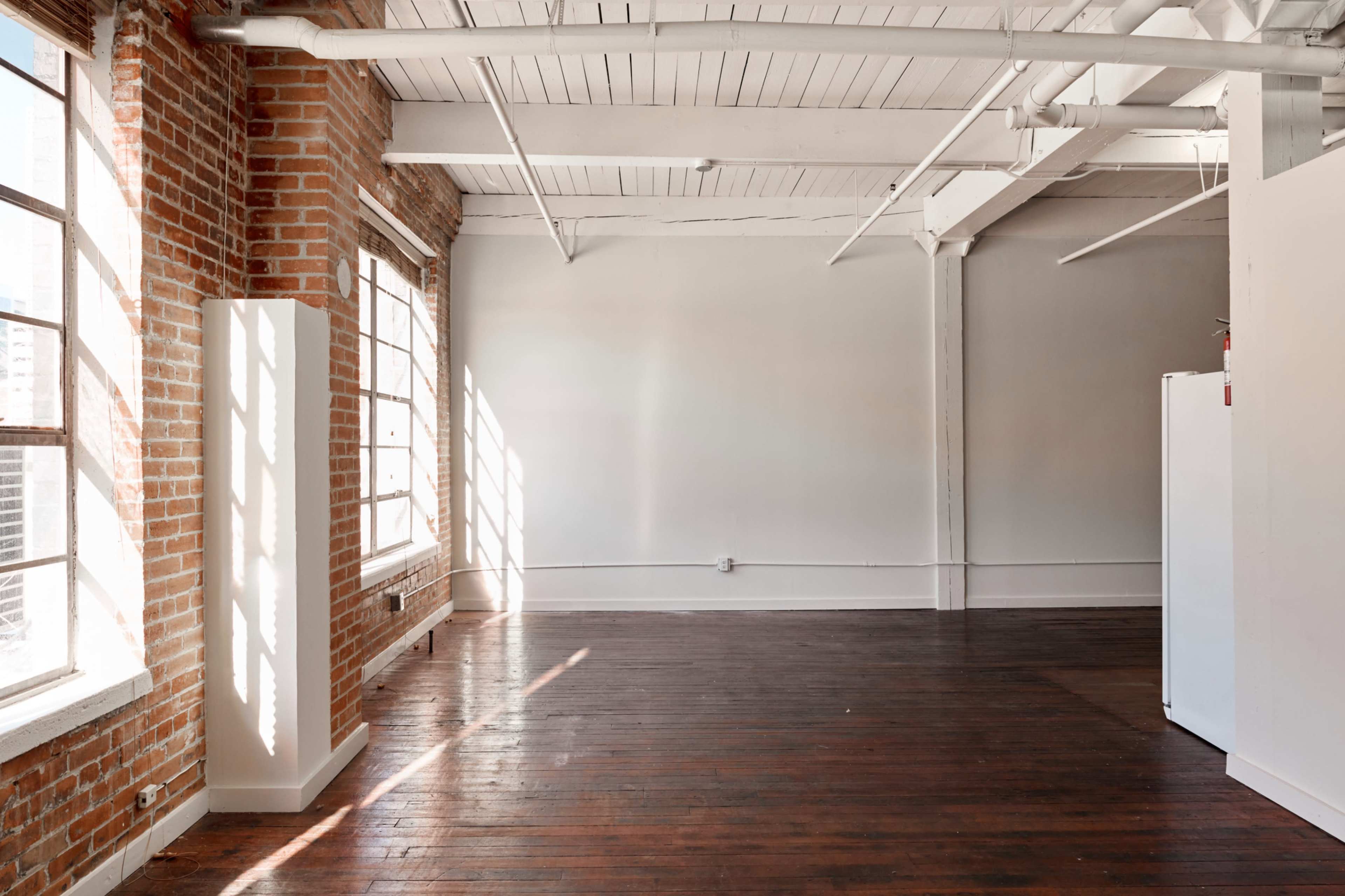 An empty room with brick walls, large windows allowing natural light, and wooden floors.
