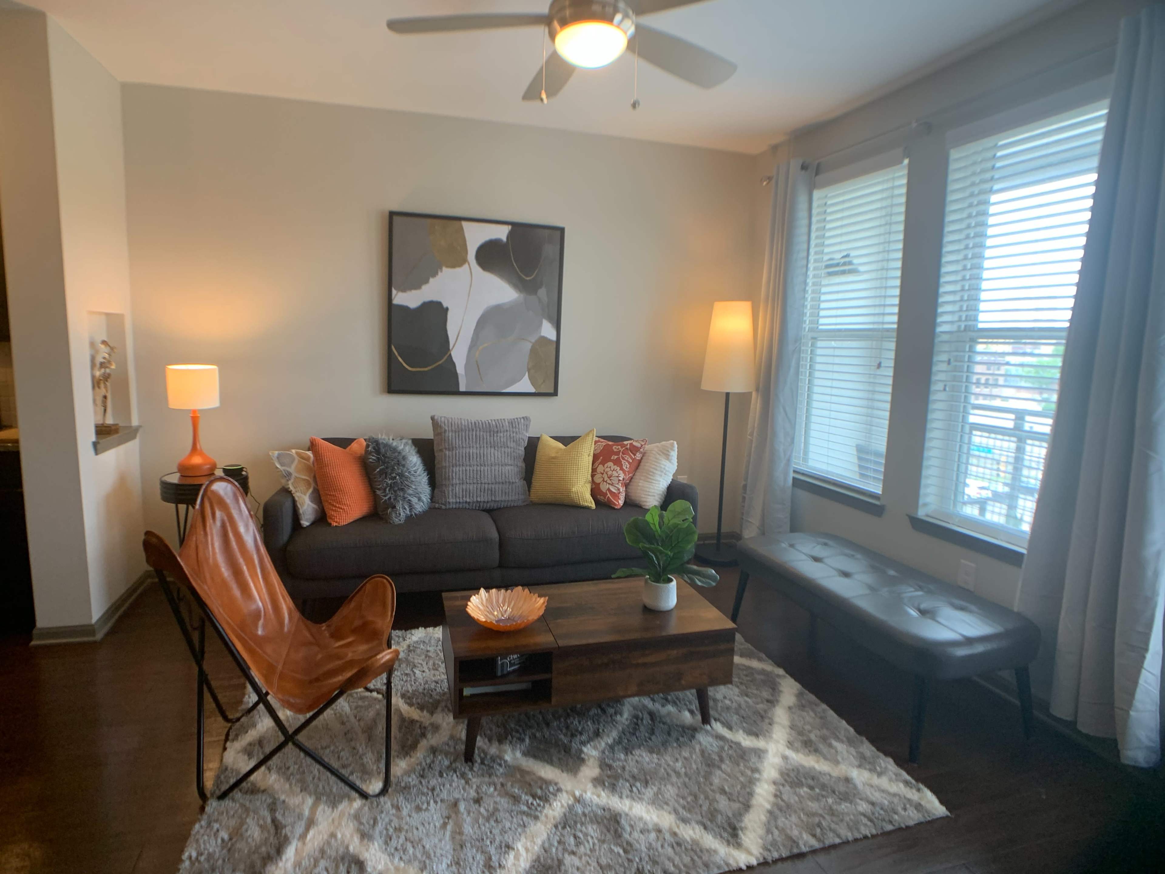 A modern living room features a gray sofa with colorful pillows, a wooden coffee table, a chair, and large windows allowing natural light to enter.