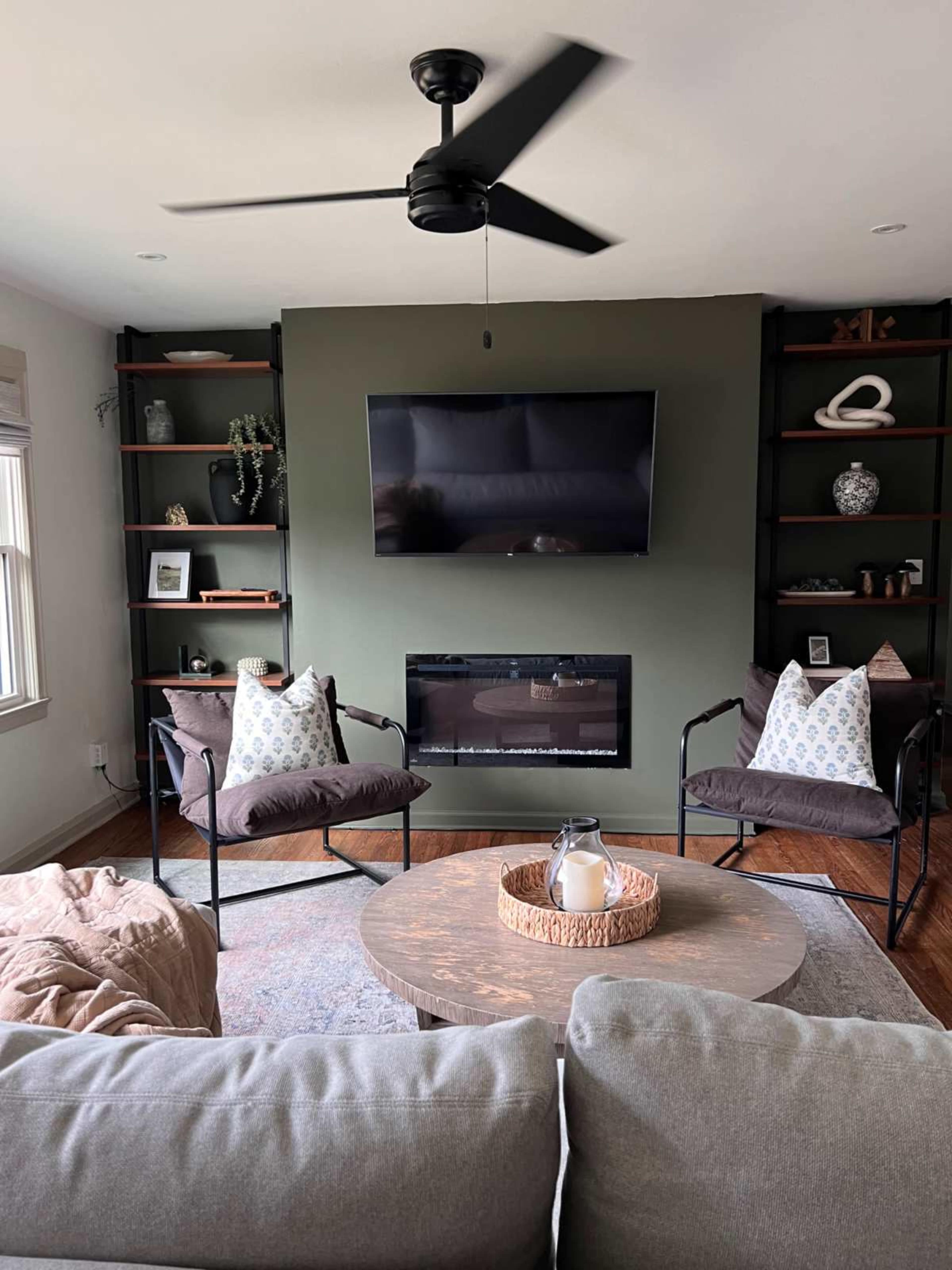 The living room features a dark green accent wall with a mounted TV above a modern fireplace, flanked by bookshelves and furnished with a circular coffee table and comfortable seating.