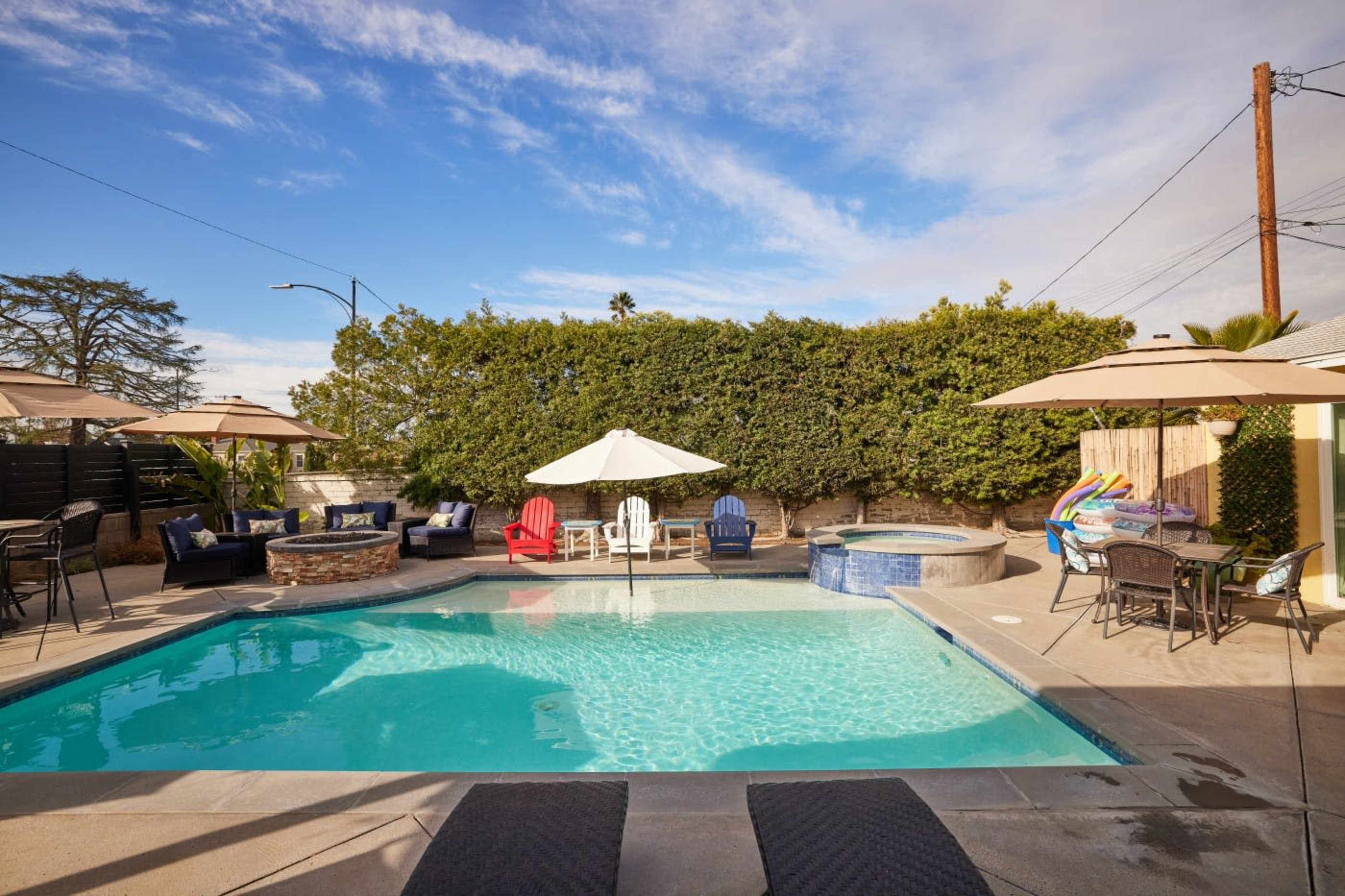 The image shows a backyard pool area featuring a swimming pool, a hot tub, lounge chairs, and several umbrellas surrounded by landscaping.