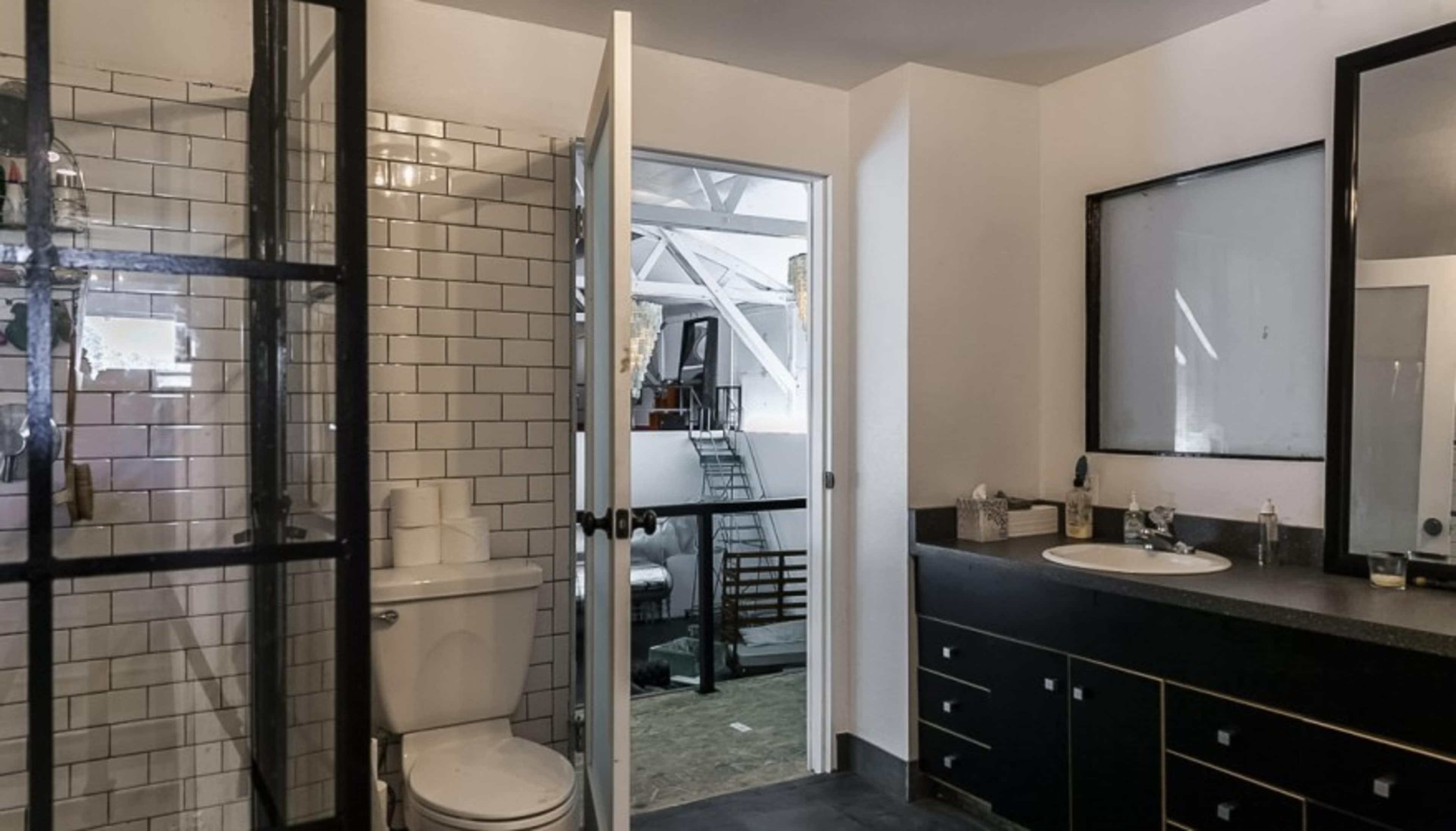 The image shows a modern bathroom featuring a toilet, a double sink vanity with dark cabinetry, and a glass door leading to a separate space.