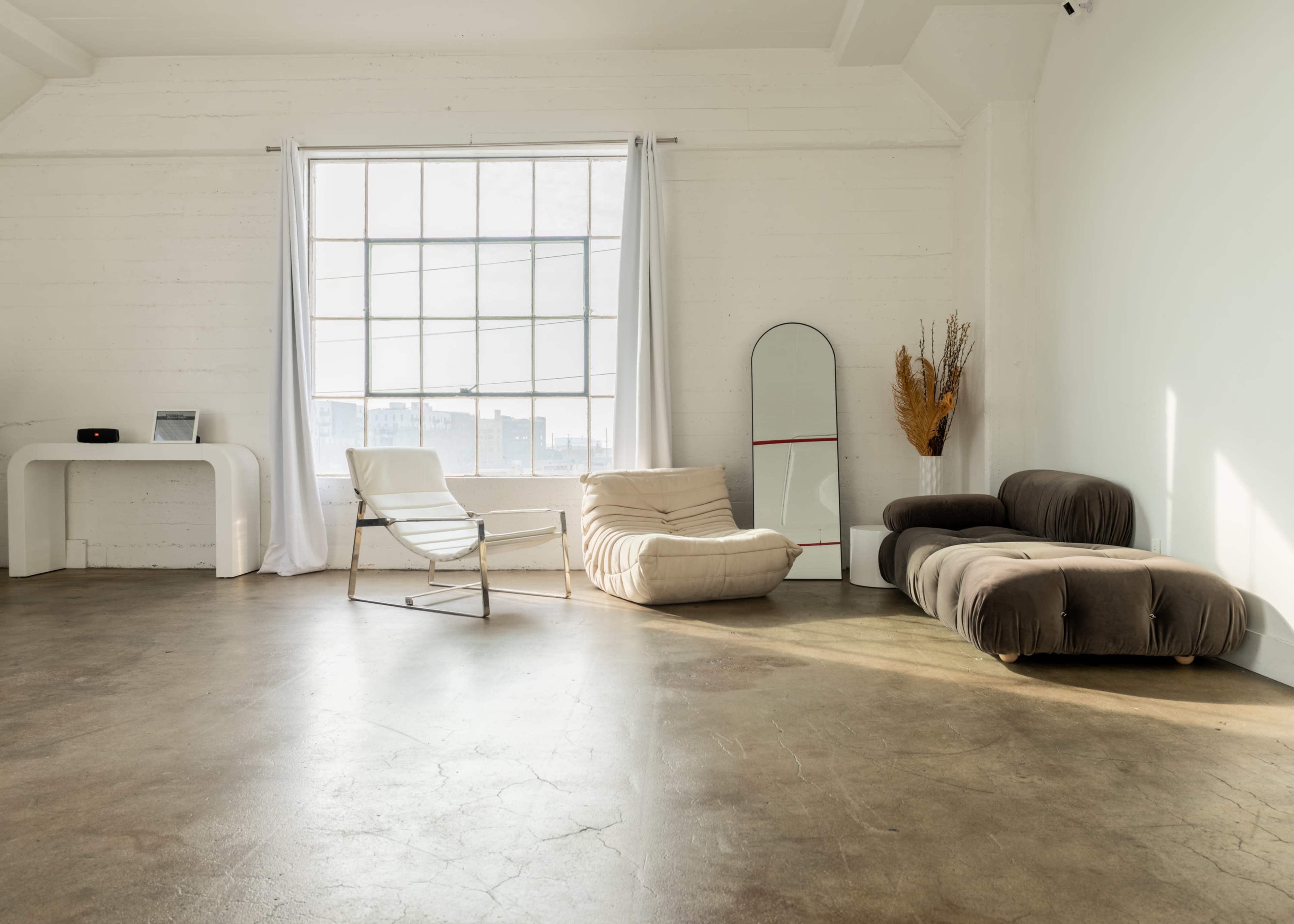 The image shows a spacious, minimalistic room with a large window, a white chair, a bean bag, a dark sofa, and a mirror against the wall.