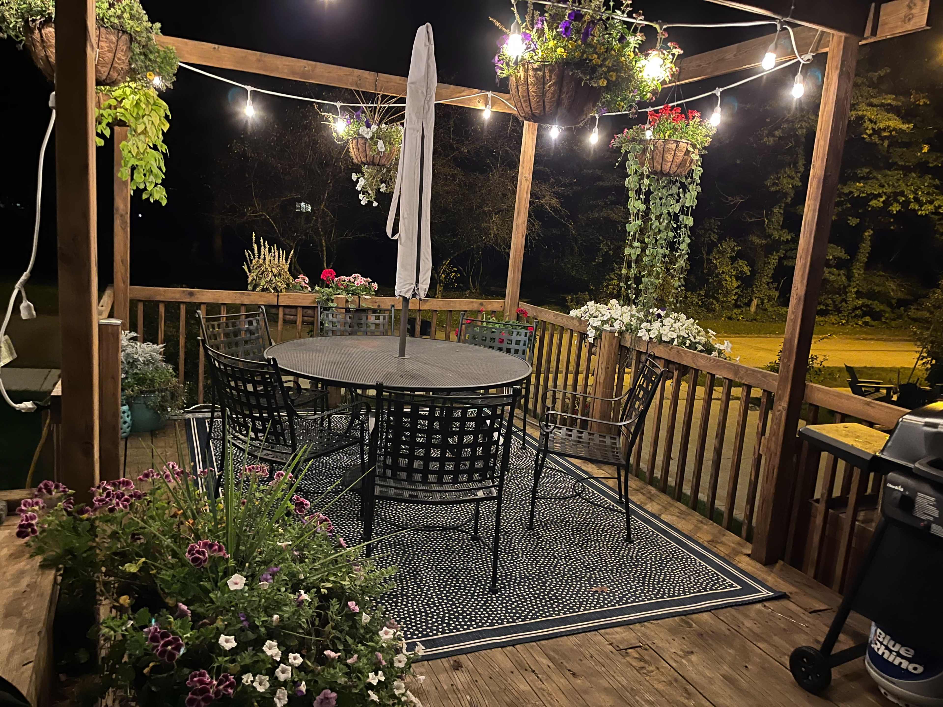 A wooden deck at night features a circular table surrounded by six black chairs, adorned with hanging planters and string lights.