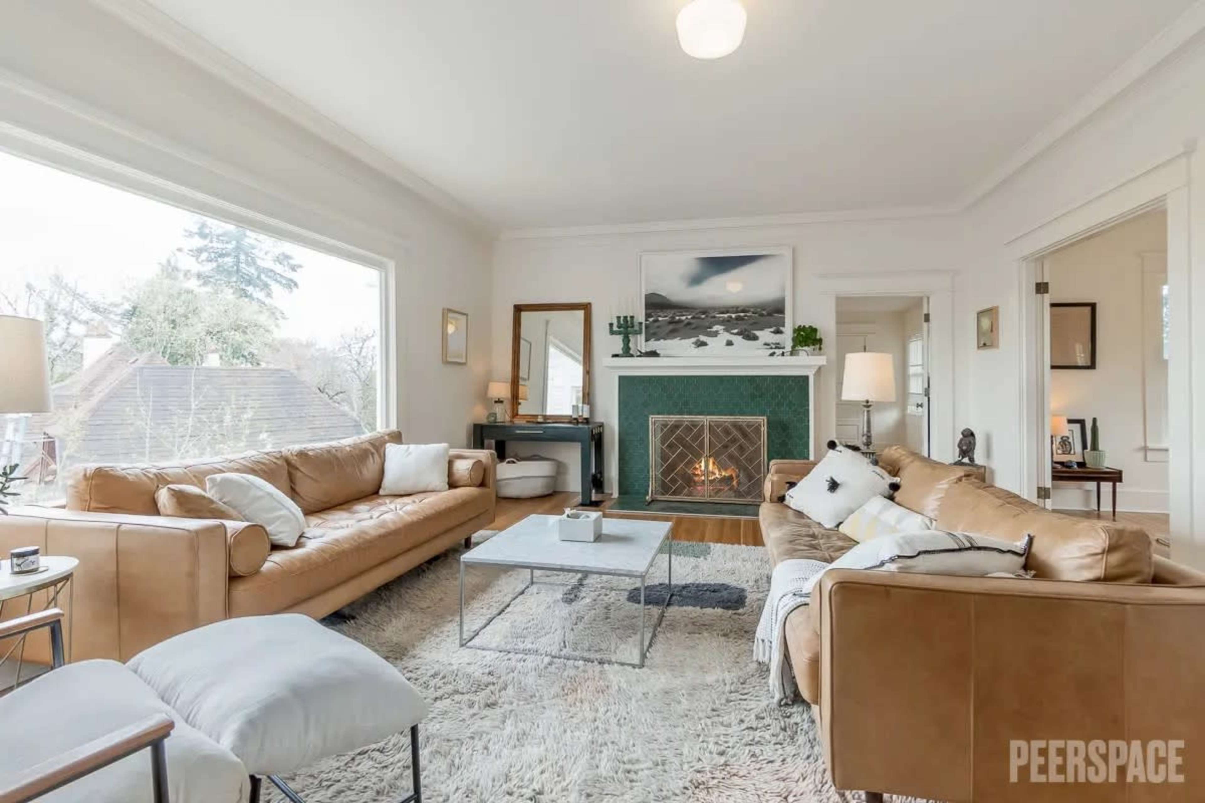 A bright living room with large windows, featuring two leather sofas facing a coffee table and a fireplace with a green tile surround.