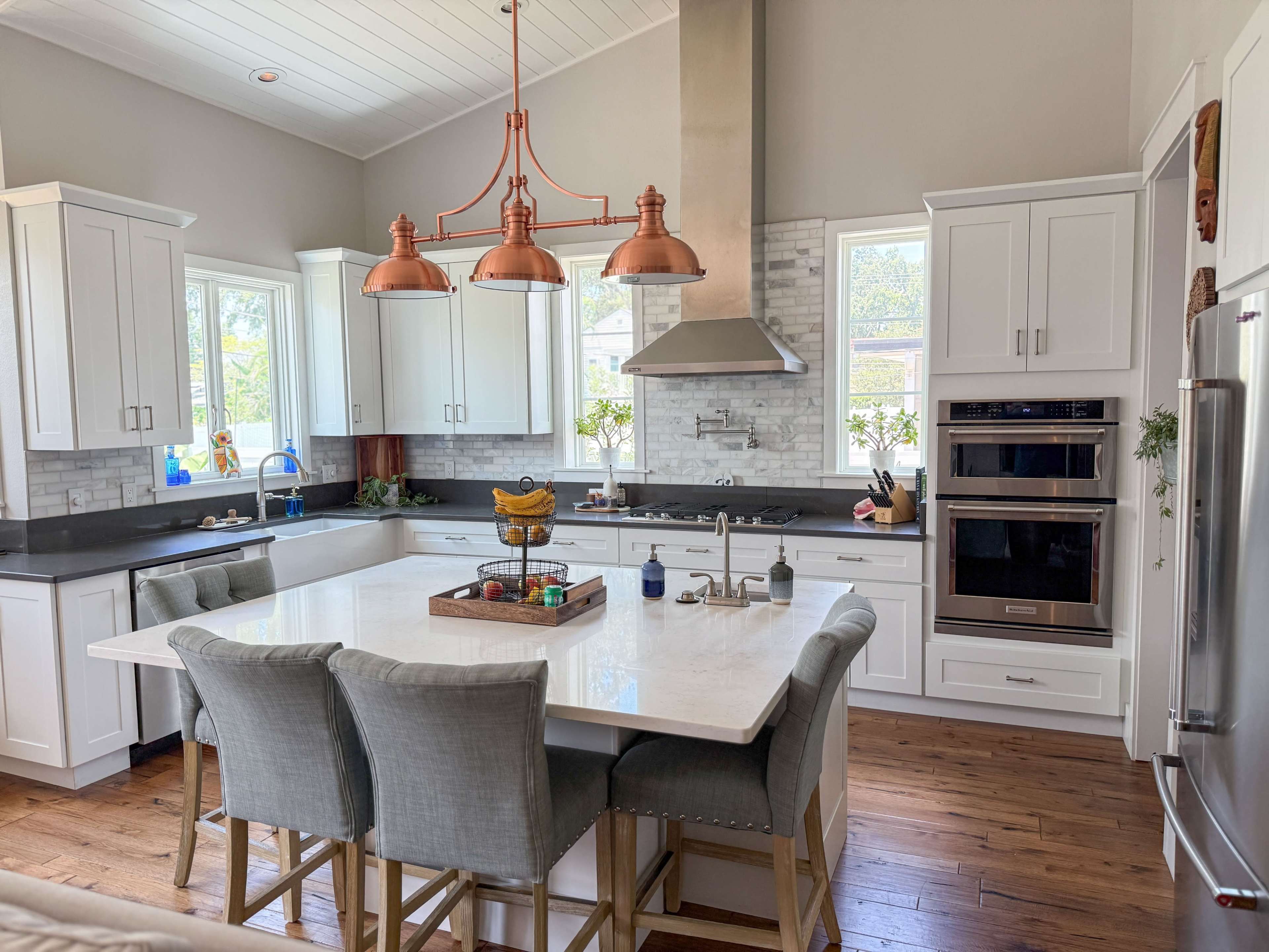 The image depicts a modern kitchen featuring white cabinets, a large island with seating, and copper pendant lights.